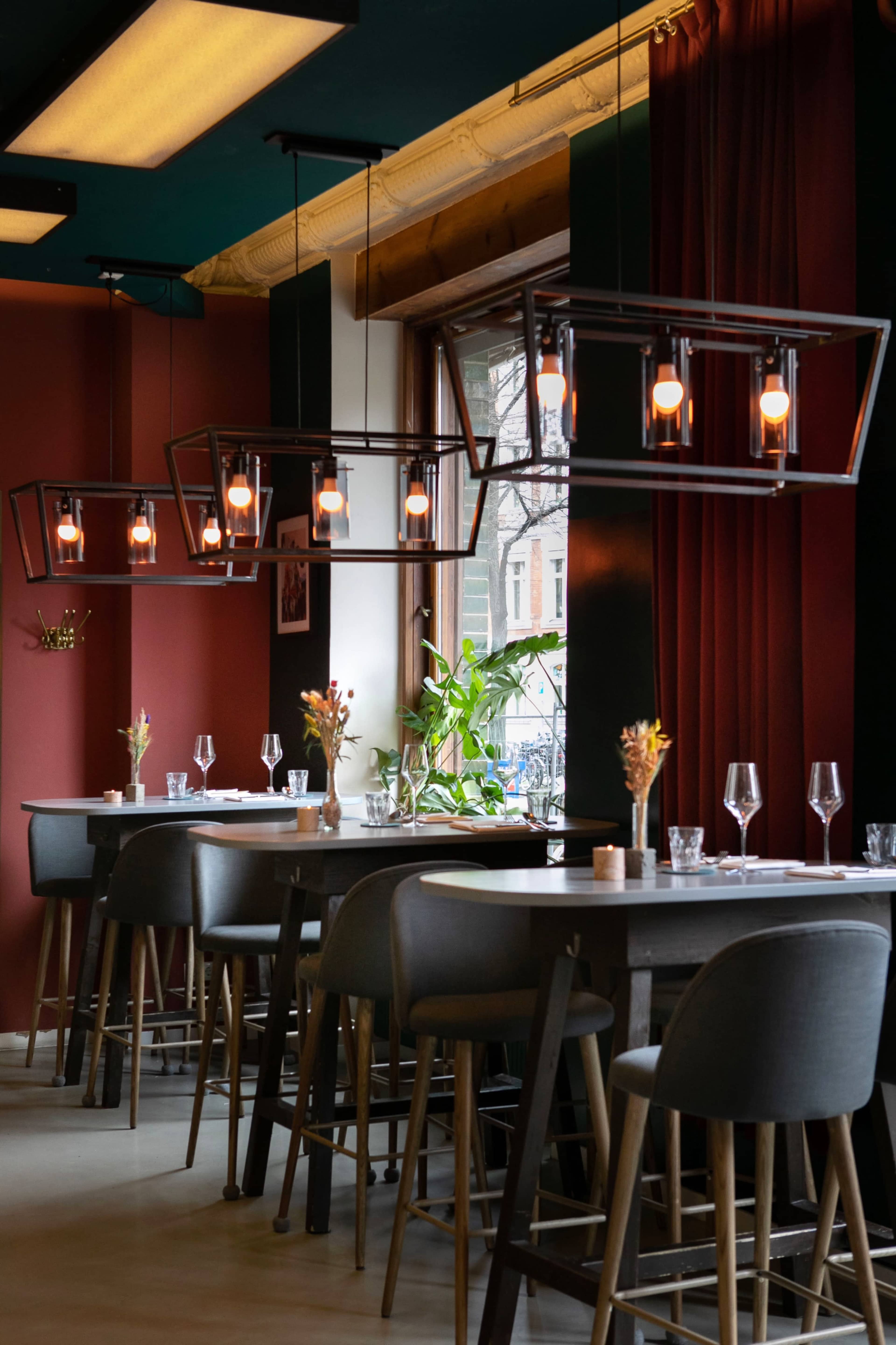 The image shows a modern restaurant interior featuring stylish pendant lighting, dark red walls, and tables set with glasses and flower arrangements.