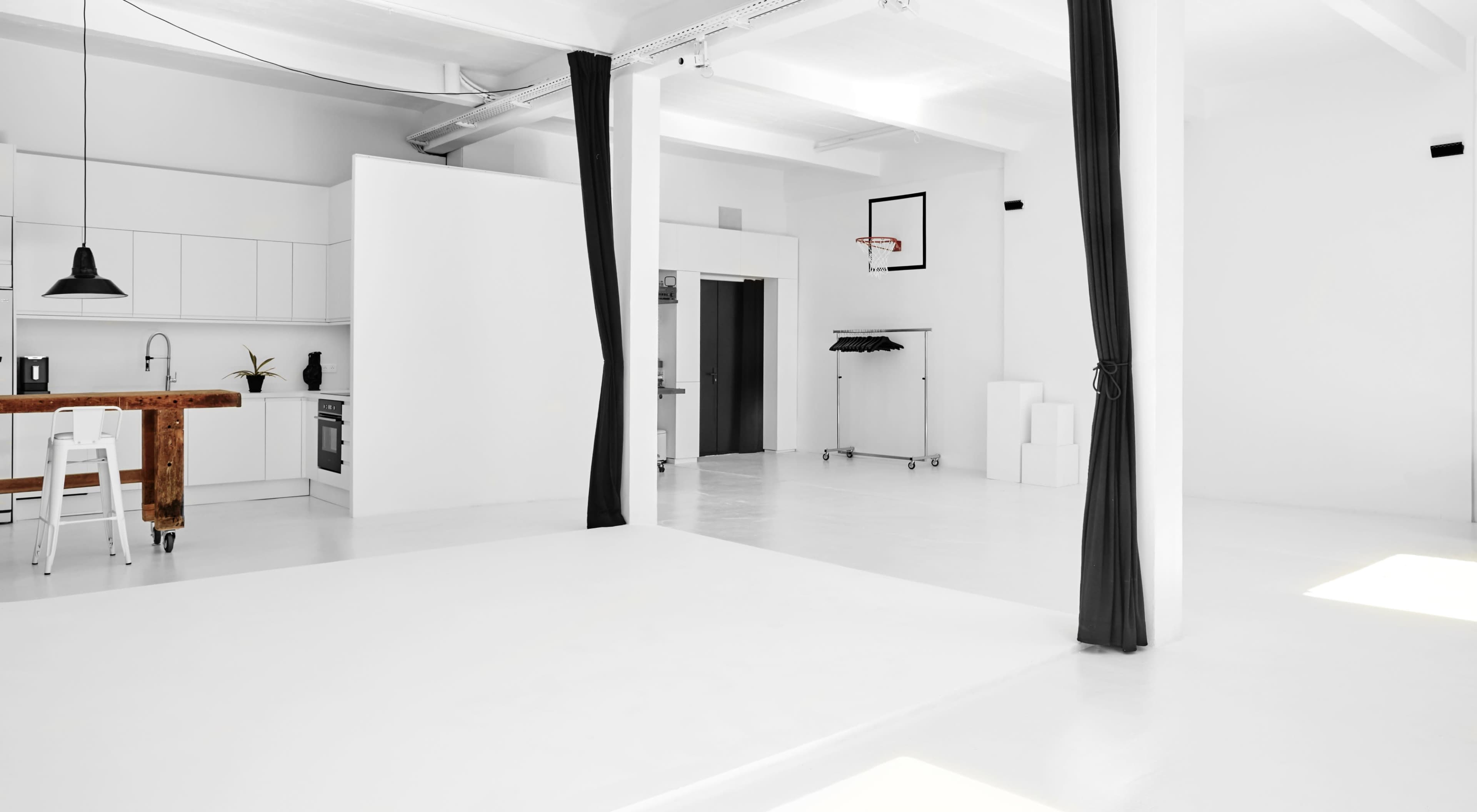 A spacious, minimalist interior with white walls and a kitchenette, featuring a basketball hoop and a clothing rack in one corner.