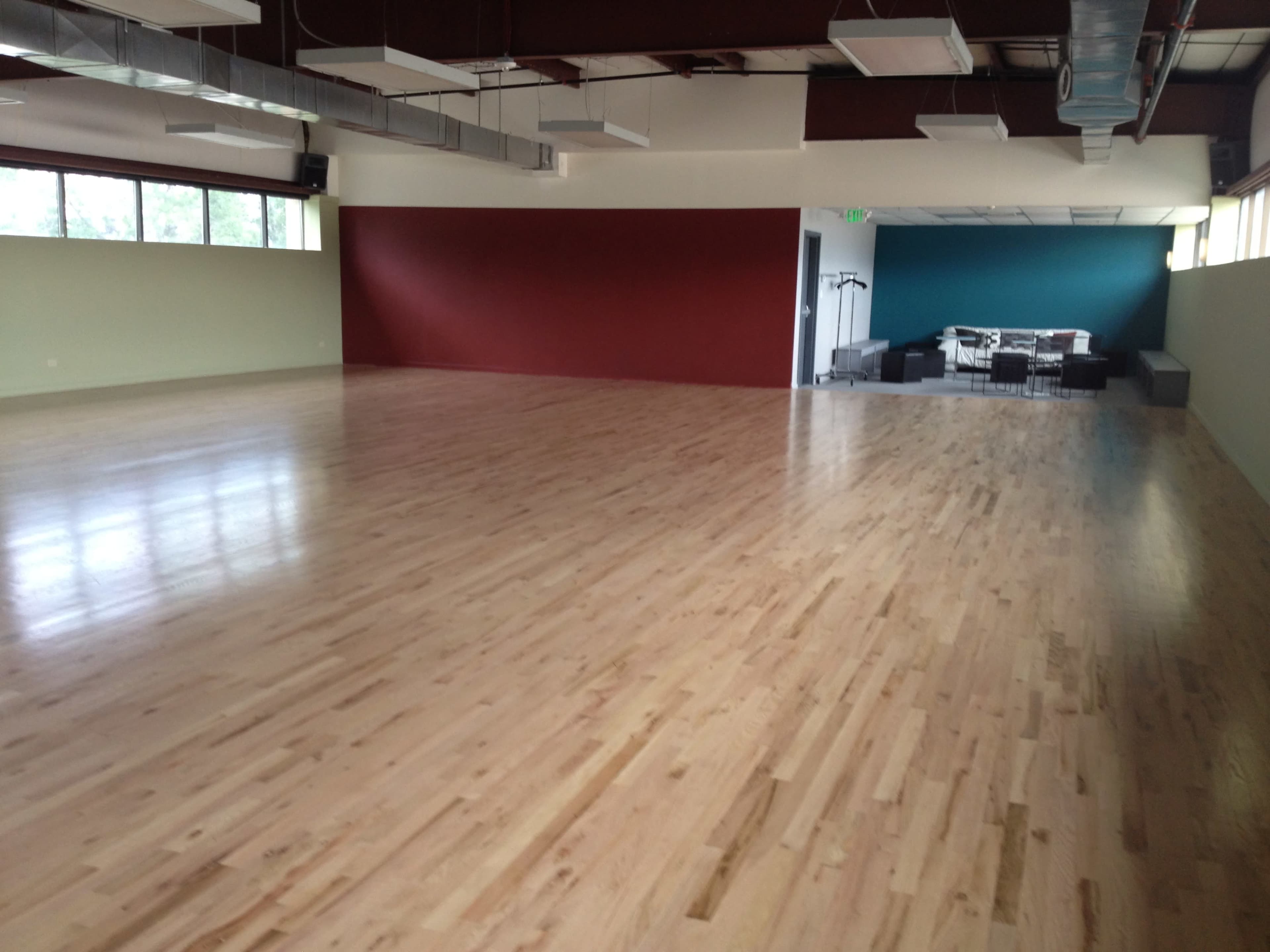 A spacious room with a polished wooden floor, large windows, and a red accent wall, leading to a partially visible area with furniture in the background.