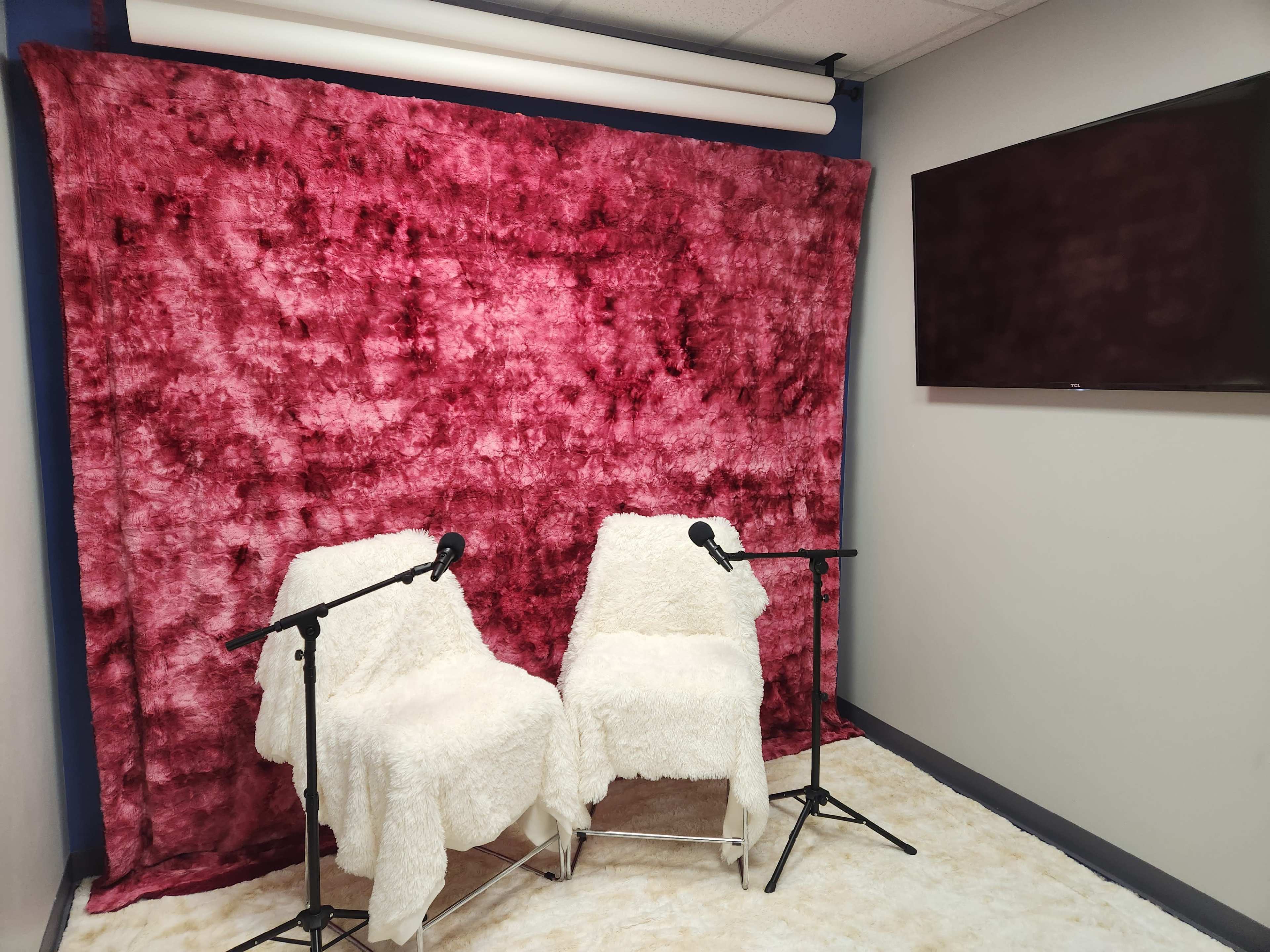 The image shows two white, fluffy chairs with microphones set up in front of a red textured backdrop and a wall-mounted TV.