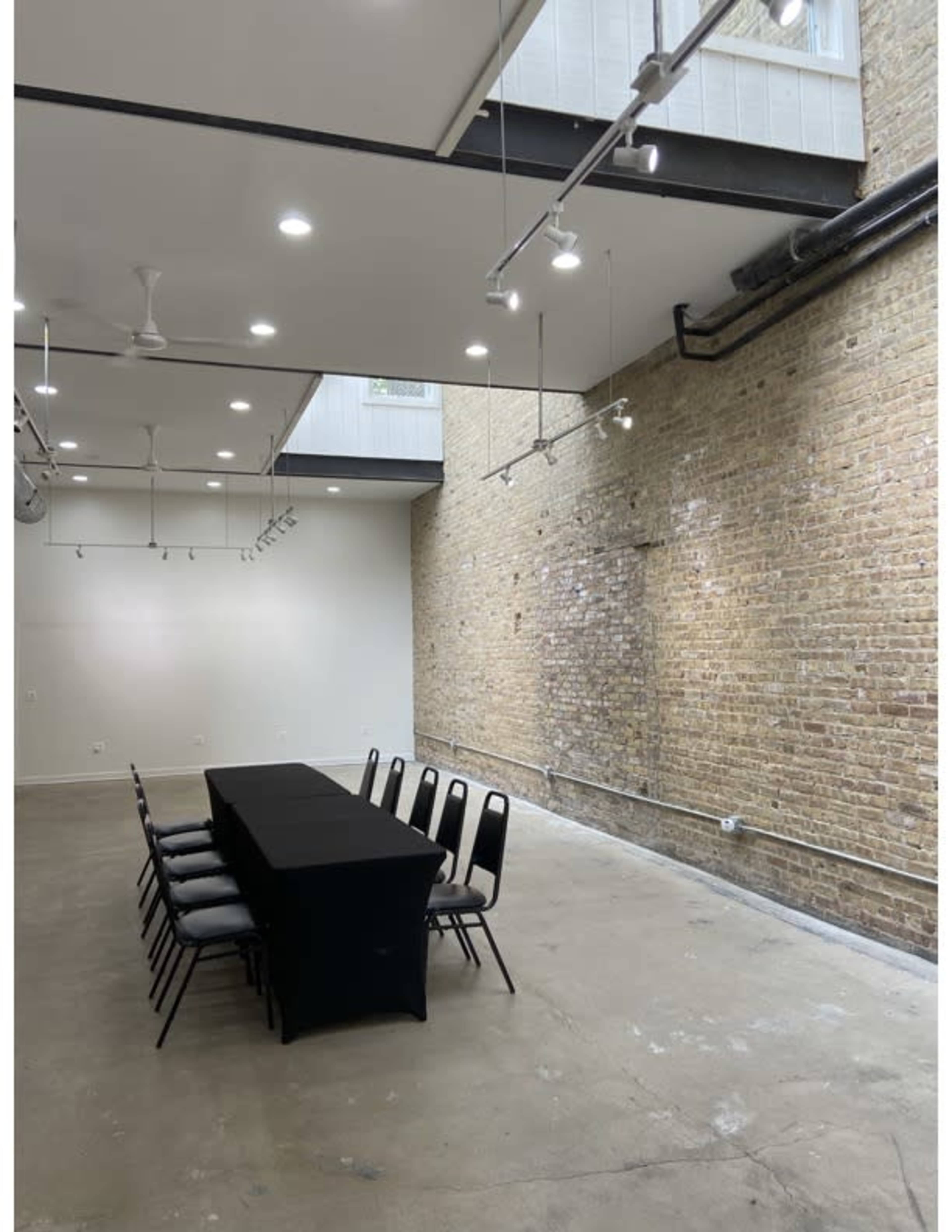A long black table with chairs is set up in an industrial-style room featuring exposed brick walls and ceiling lights.