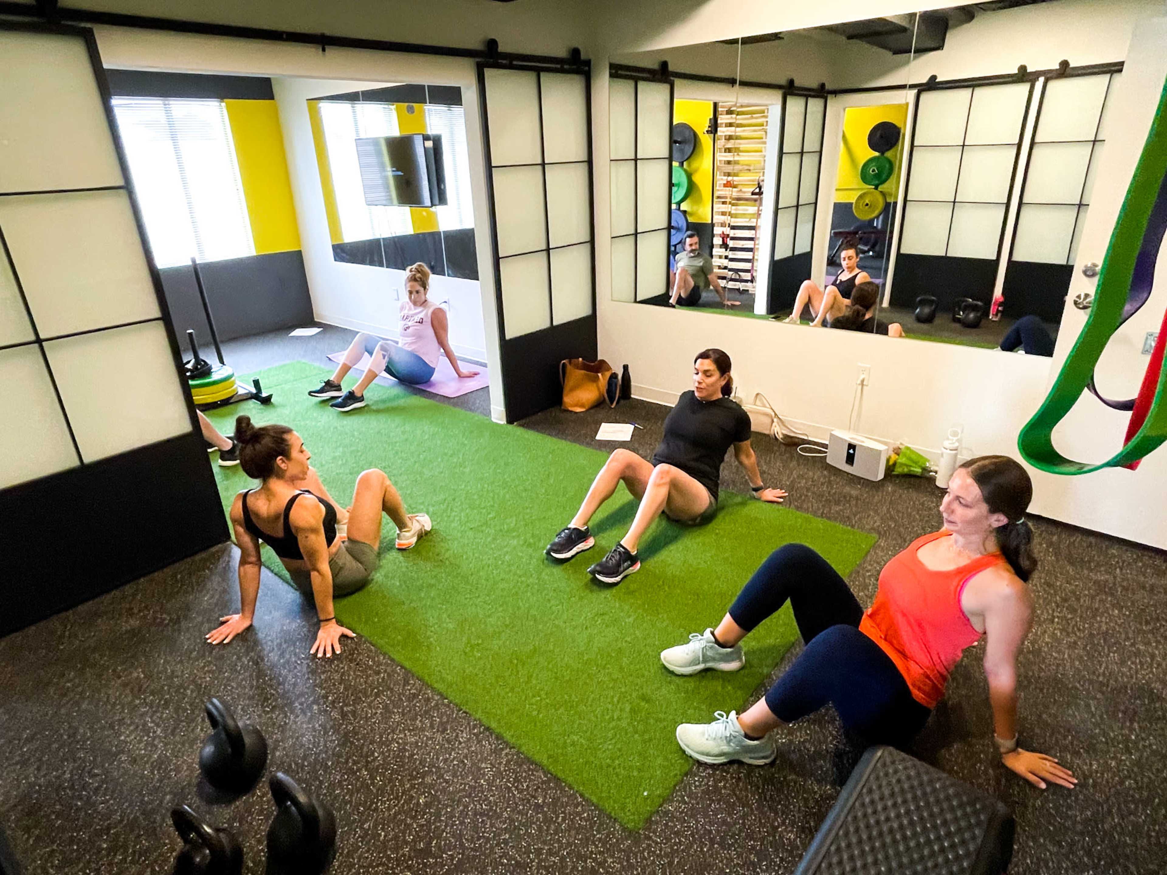 Three women are seated on a green exercise mat in a gym, while a fourth woman exercises in the background.