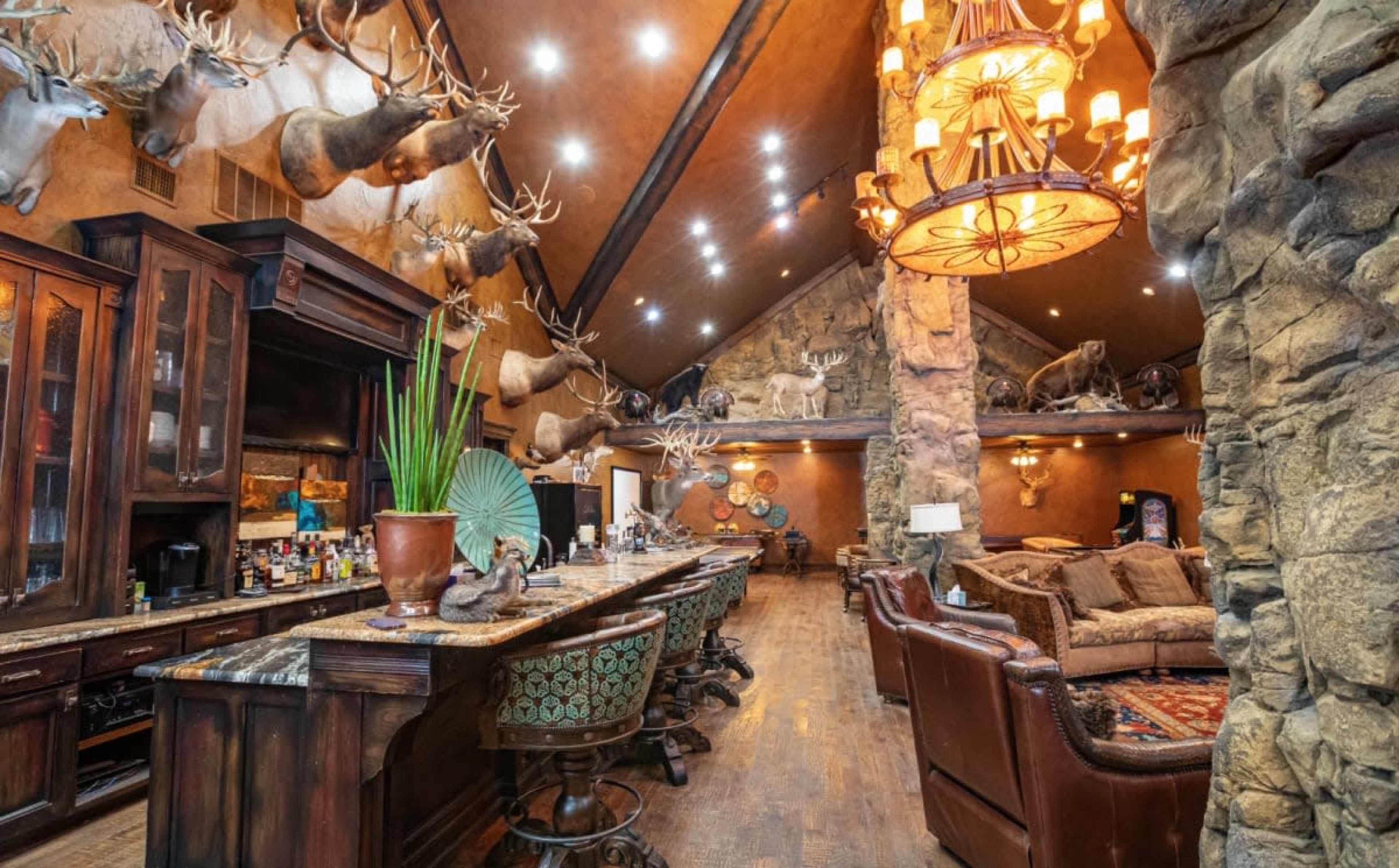 The image shows a spacious, rustic bar area featuring stone walls, a large wooden counter, and numerous mounted animal heads on display, with a high ceiling and elegant light fixtures.