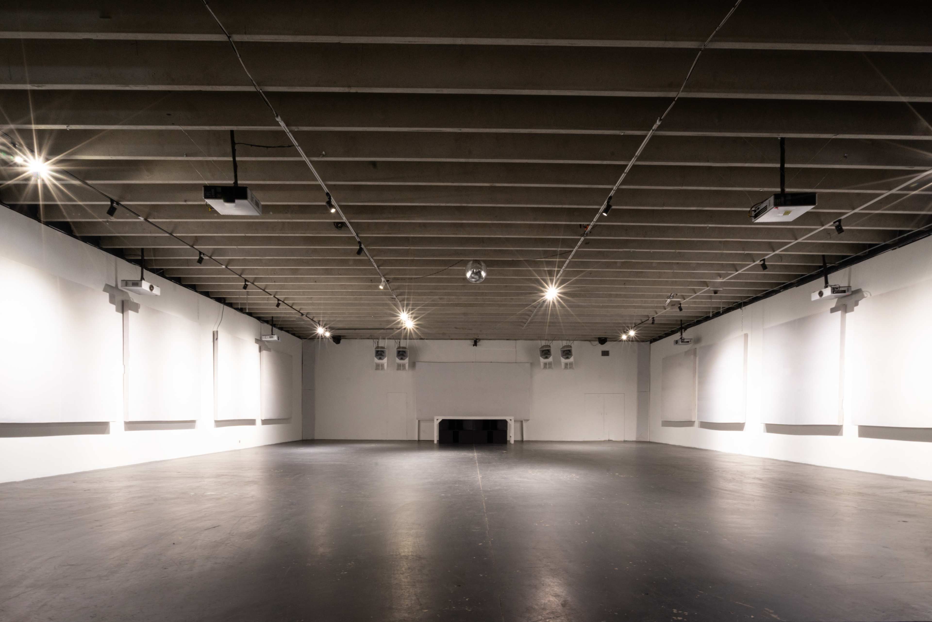The image shows a spacious, empty room with high ceilings, polished black flooring, and projector equipment mounted on the walls.
