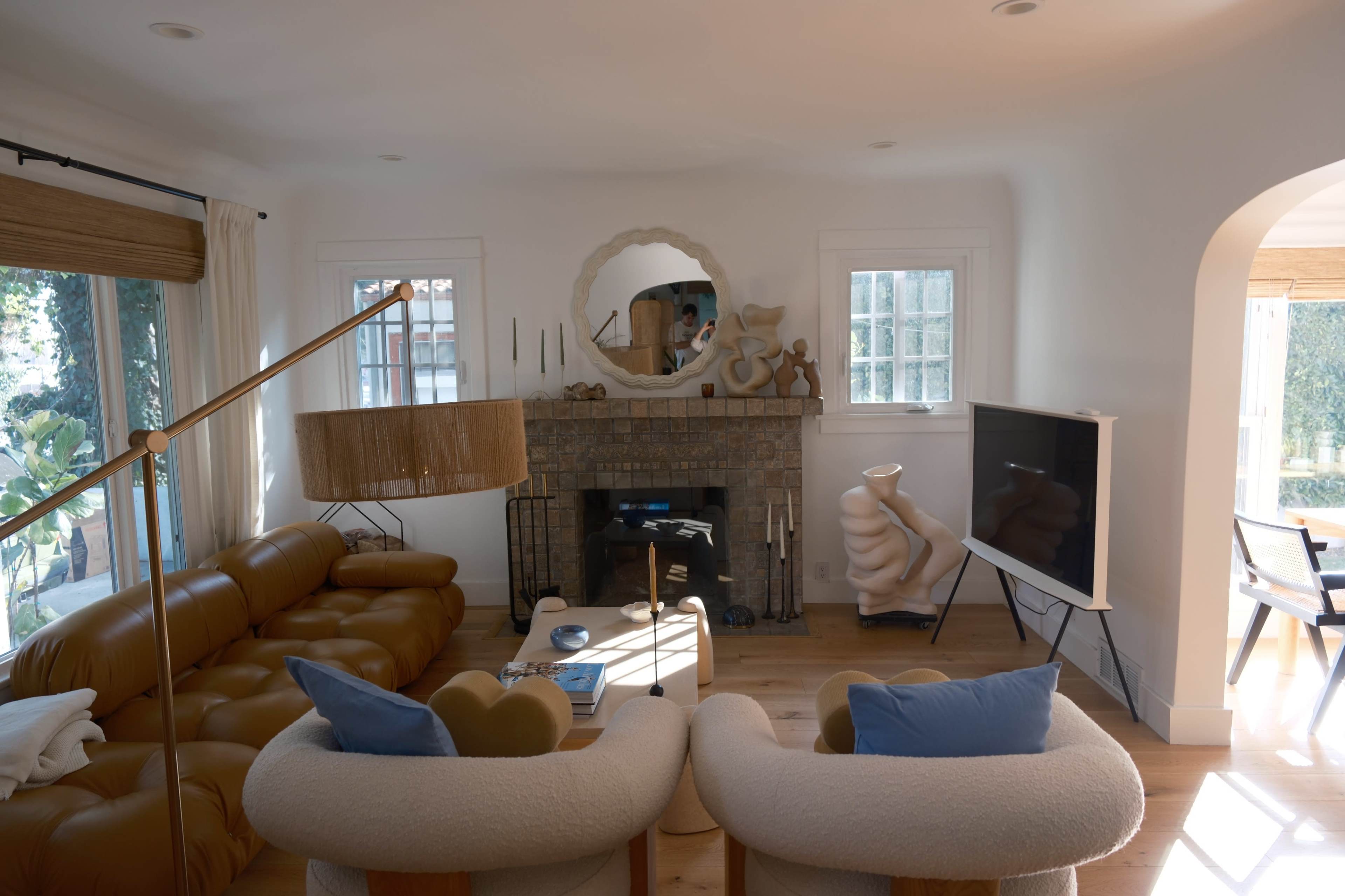 A living room features a brown leather sofa, two white armchairs, a circular mirror above a fireplace, and a television mounted on a white stand.