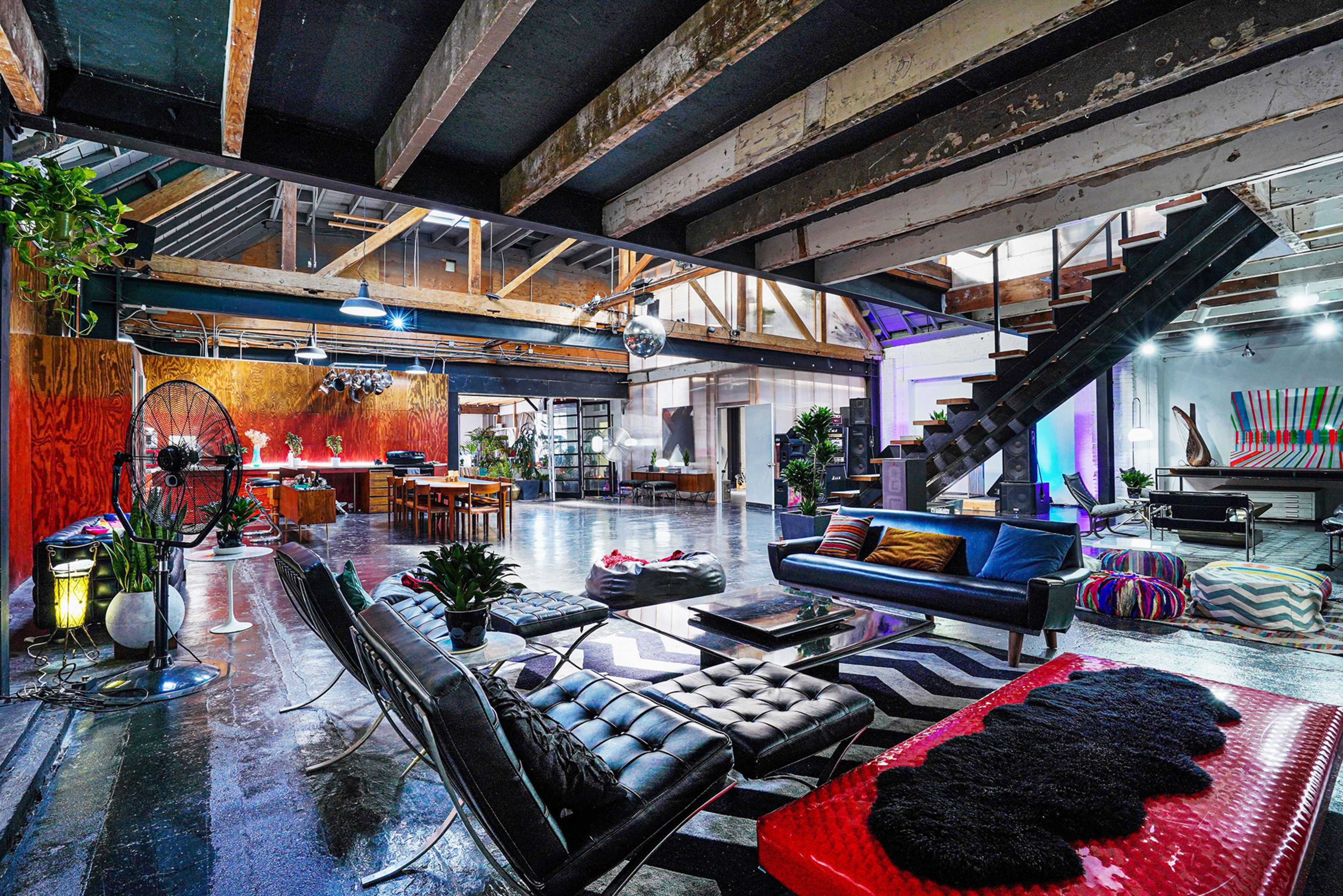 A spacious, modern loft with high ceilings, exposed beams, a mix of seating areas, and a staircase leading to an upper level.