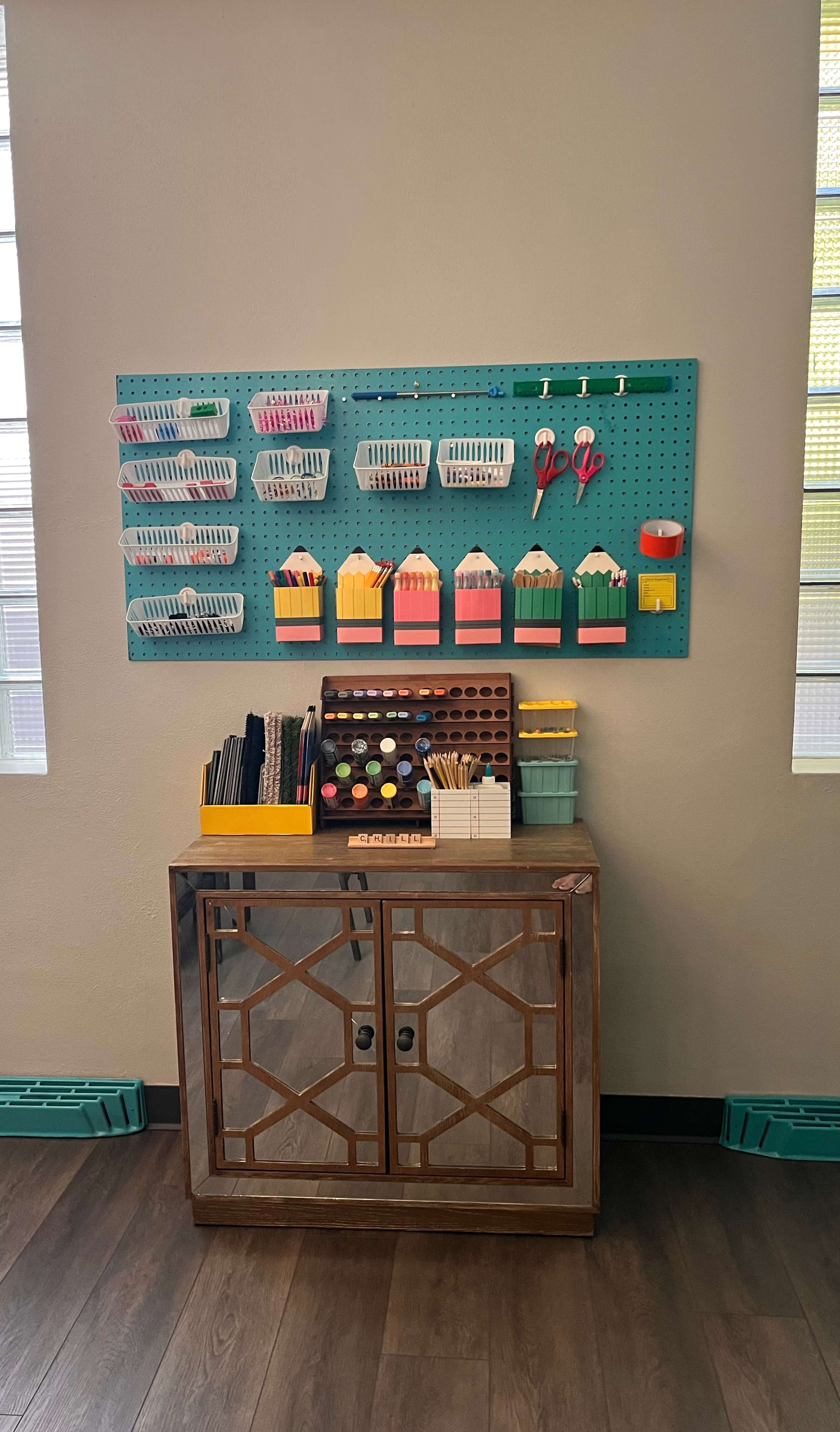A wall-mounted pegboard displays various organizational containers and tools above a wooden cabinet with a mirrored front, creating an organized workspace in a well-lit room.