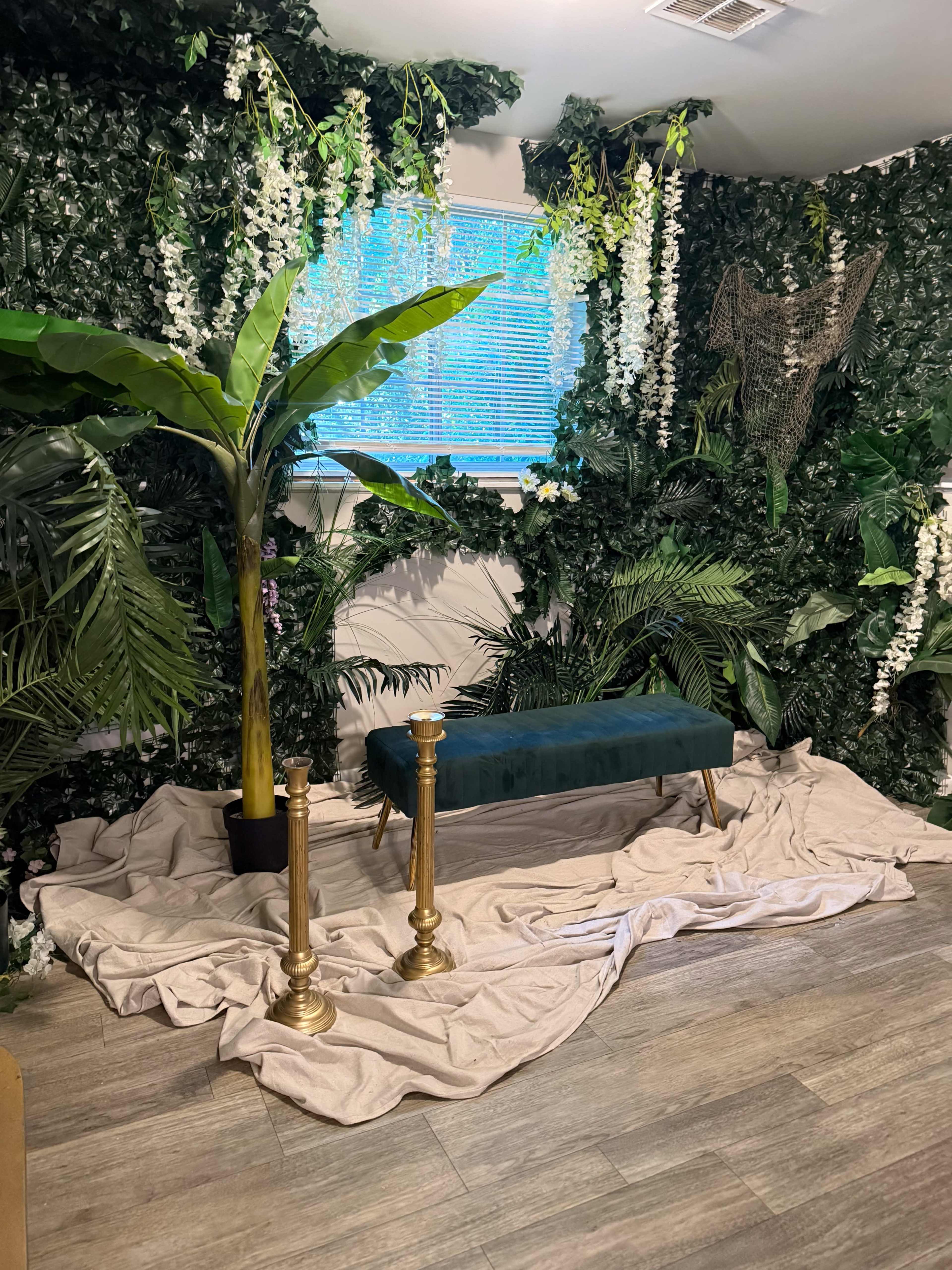 The image shows a decorated indoor space featuring lush greenery, a plush teal bench, and golden stands, all arranged over a draped light-colored fabric.