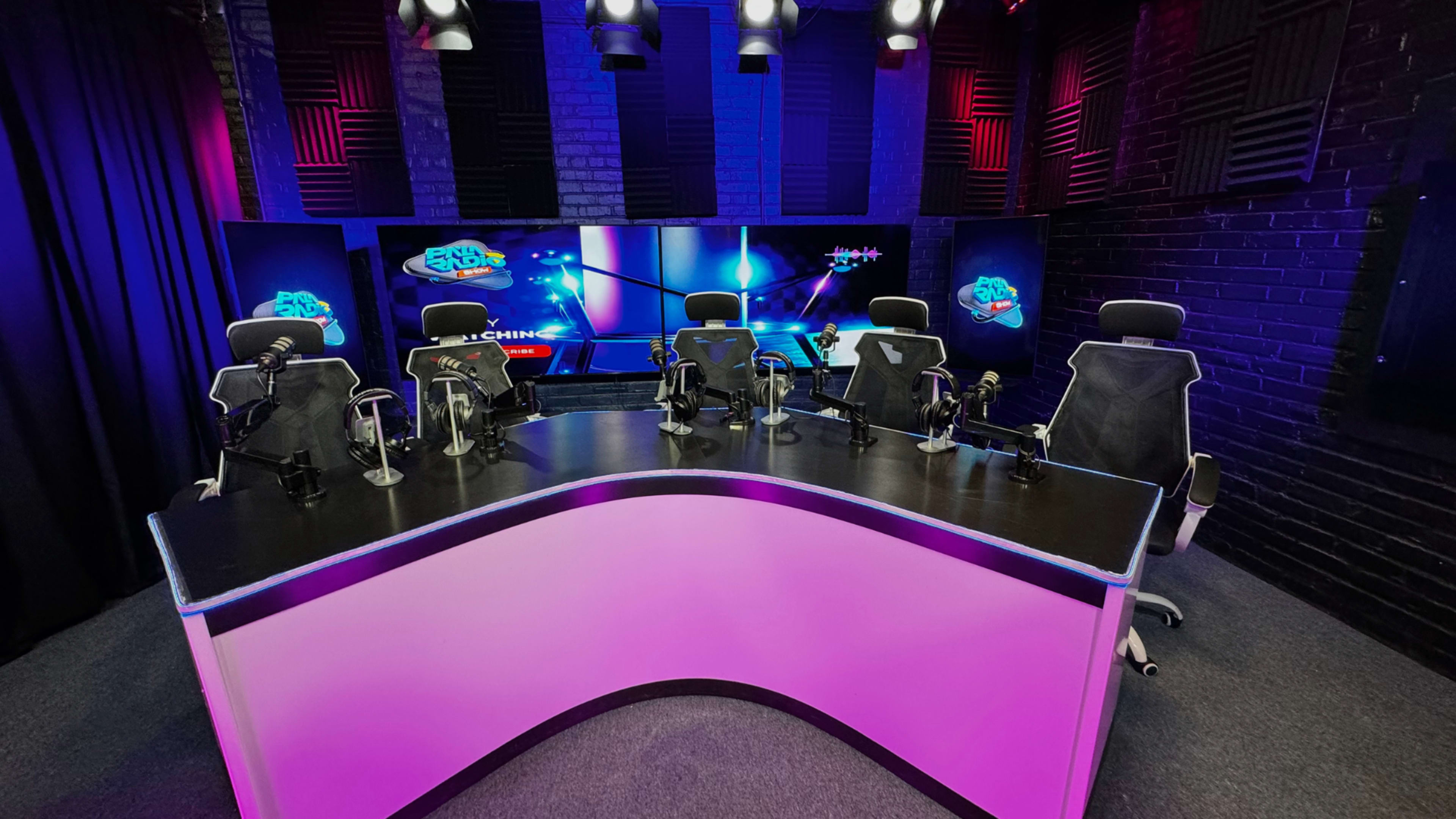 A modern studio setup with a central curved desk and several empty gaming chairs, illuminated by colorful lights and featuring large screens in the background.