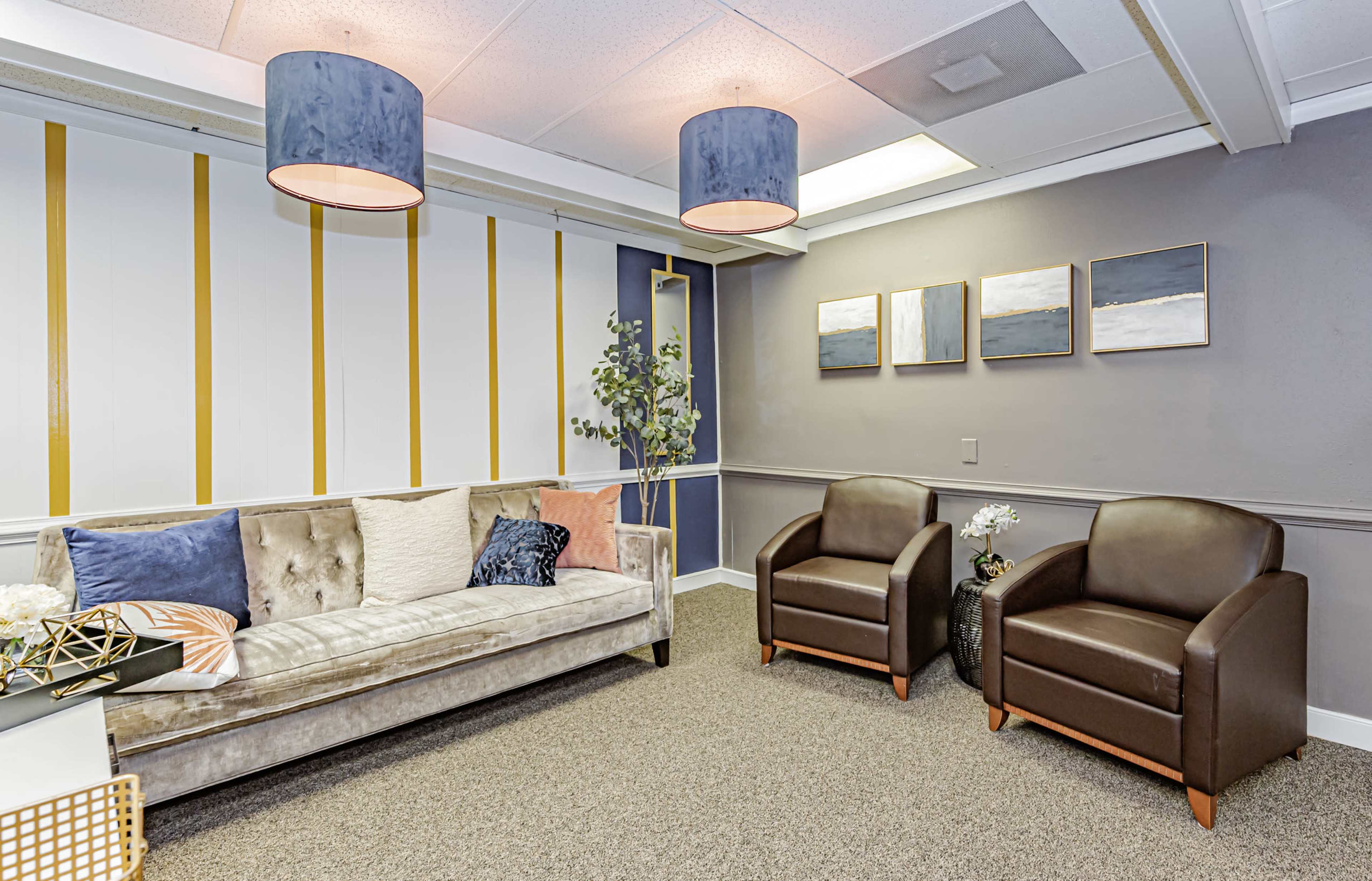 A modern lounge area with a gray sofa, two brown chairs, and abstract artworks on the wall, illuminated by pendant lighting.