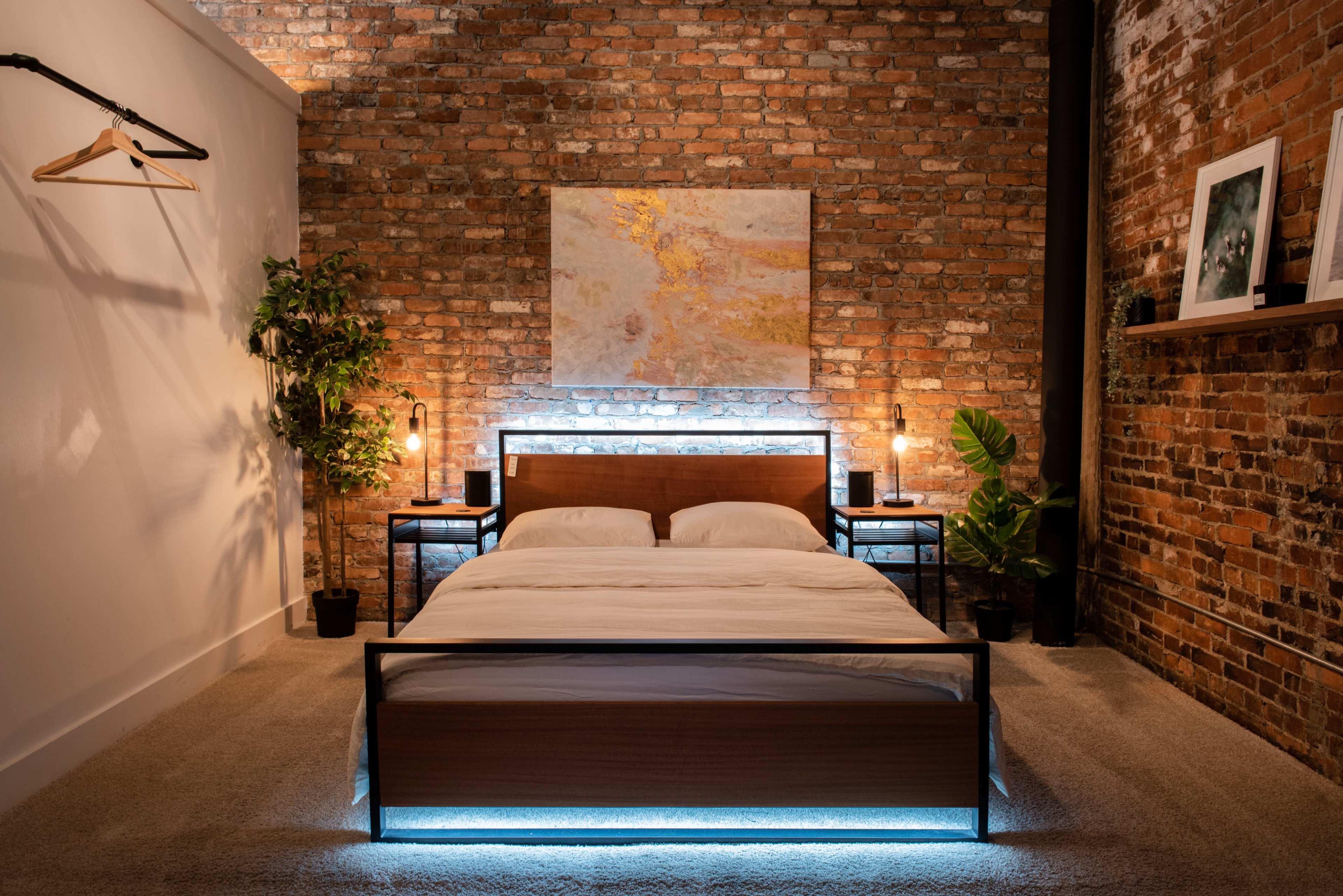 A modern bedroom featuring a wooden bed with a lighted frame, set against a brick wall, with bedside tables and decorative plants.