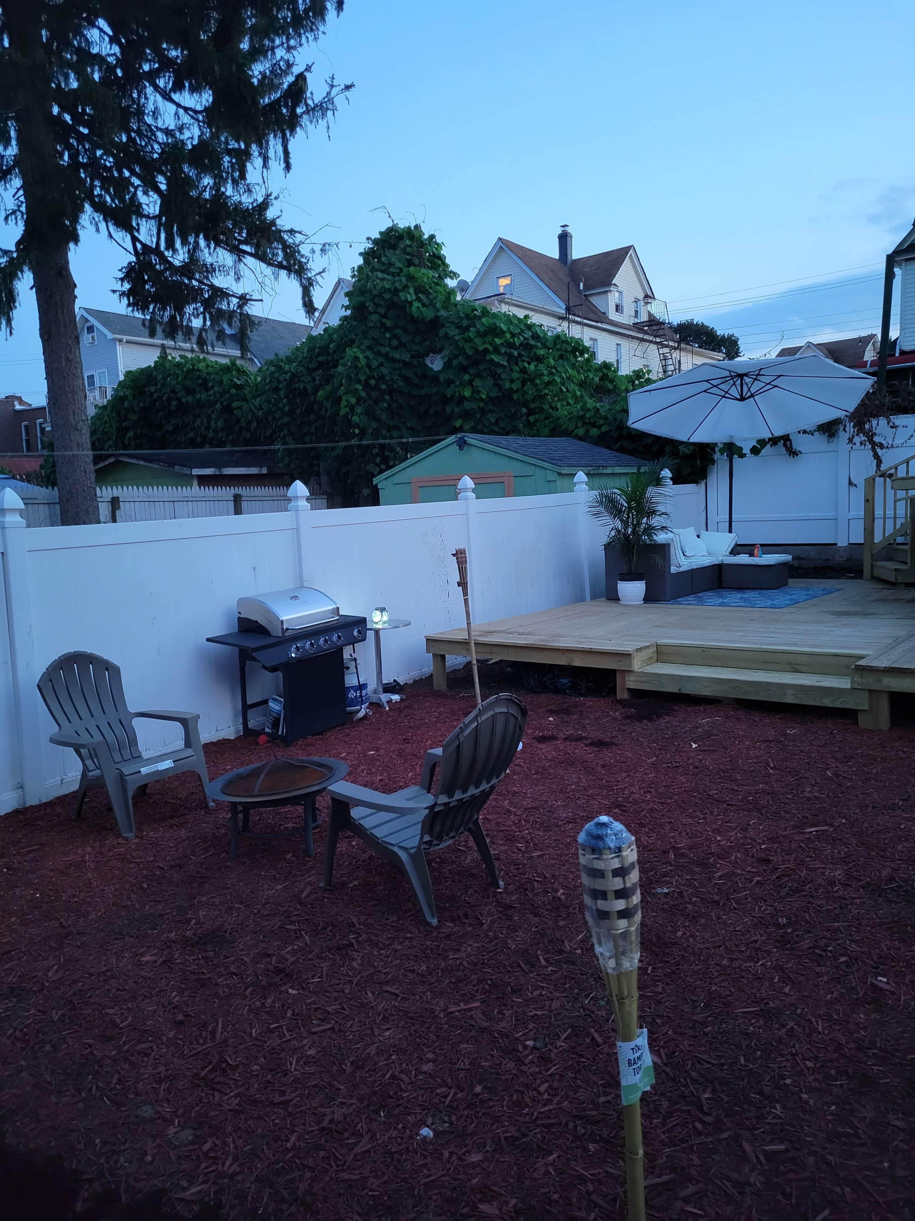 A backyard area with a grill, two chairs around a small fire pit, and a deck with an umbrella and a nearby storage shed.