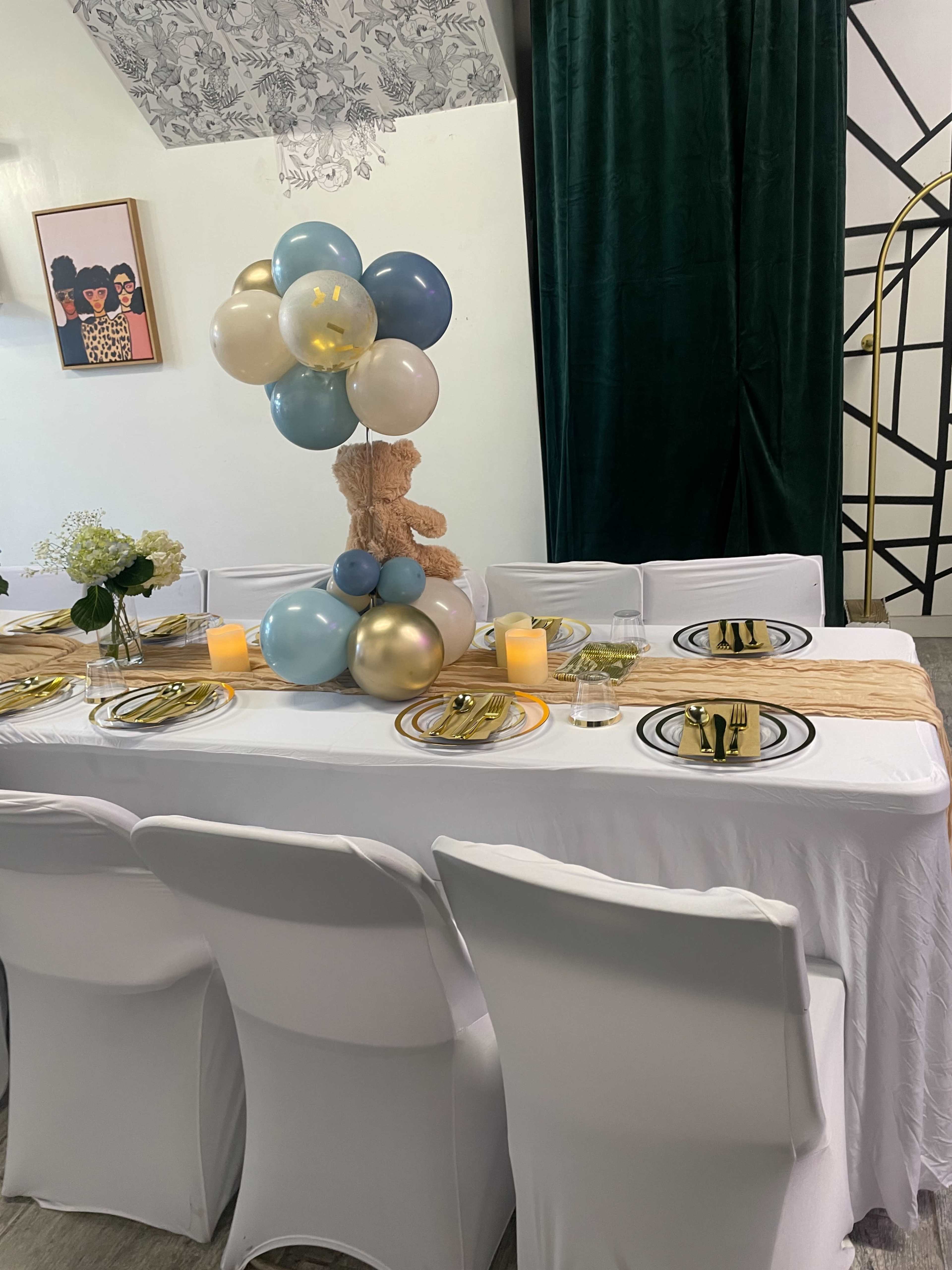 A dining table is elegantly set with white tablecloths, gold utensils, decorative plates, and a centerpiece of balloons and a teddy bear.