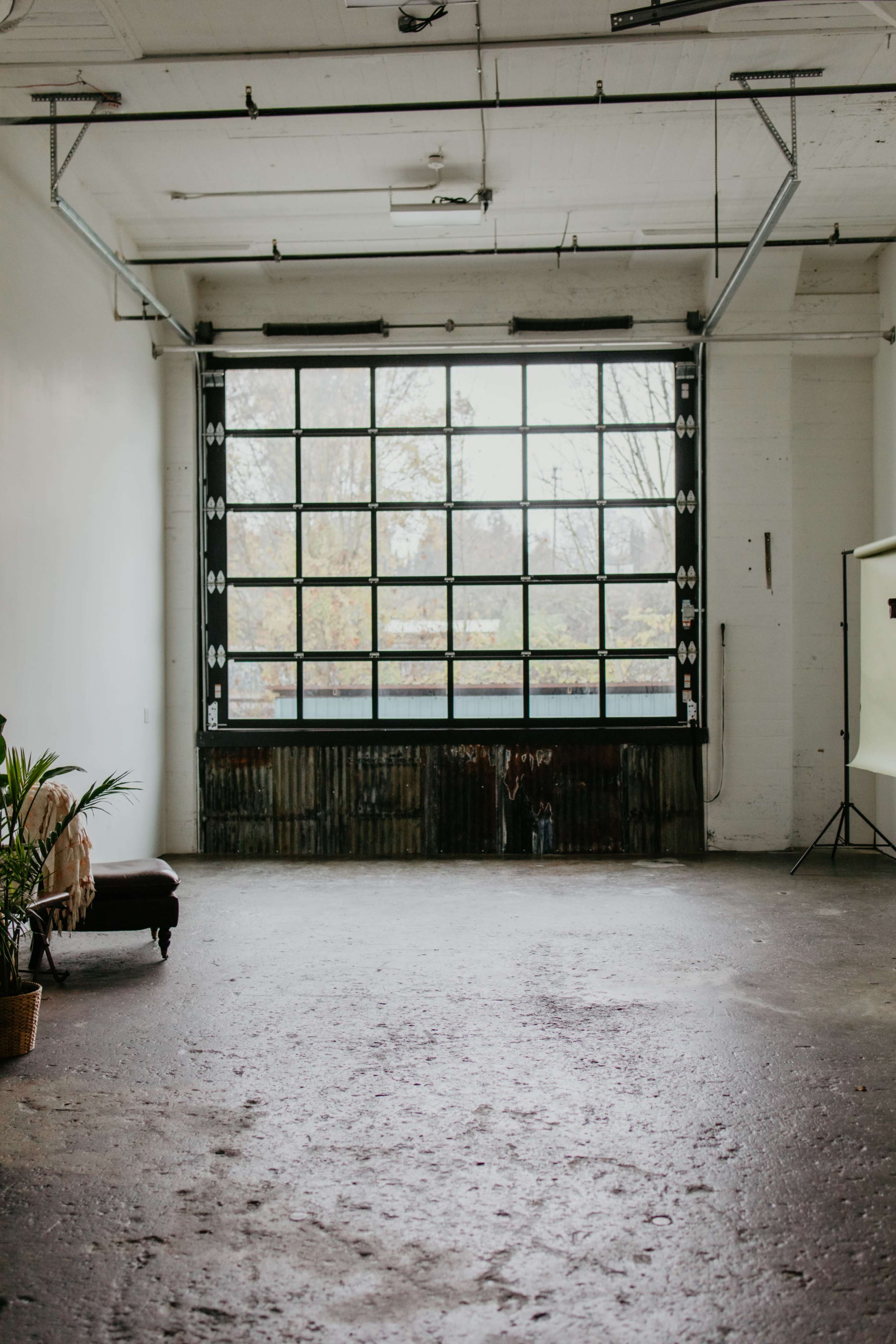The image shows a spacious, industrial room with a large window, concrete floor, and a few pieces of furniture, including a couch and a plant.