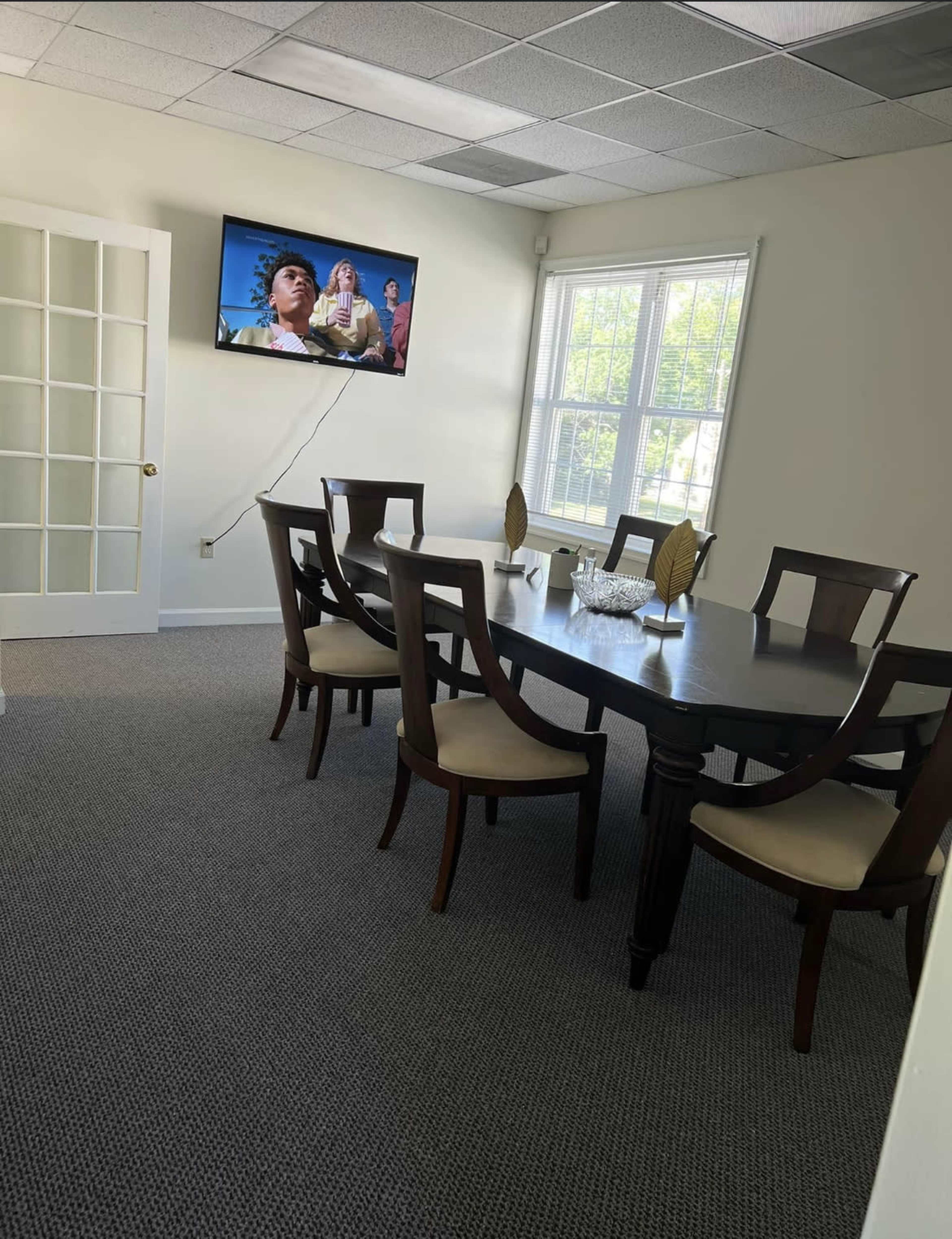 A dining room features a large table surrounded by chairs, with a television mounted on the wall showing a movie.