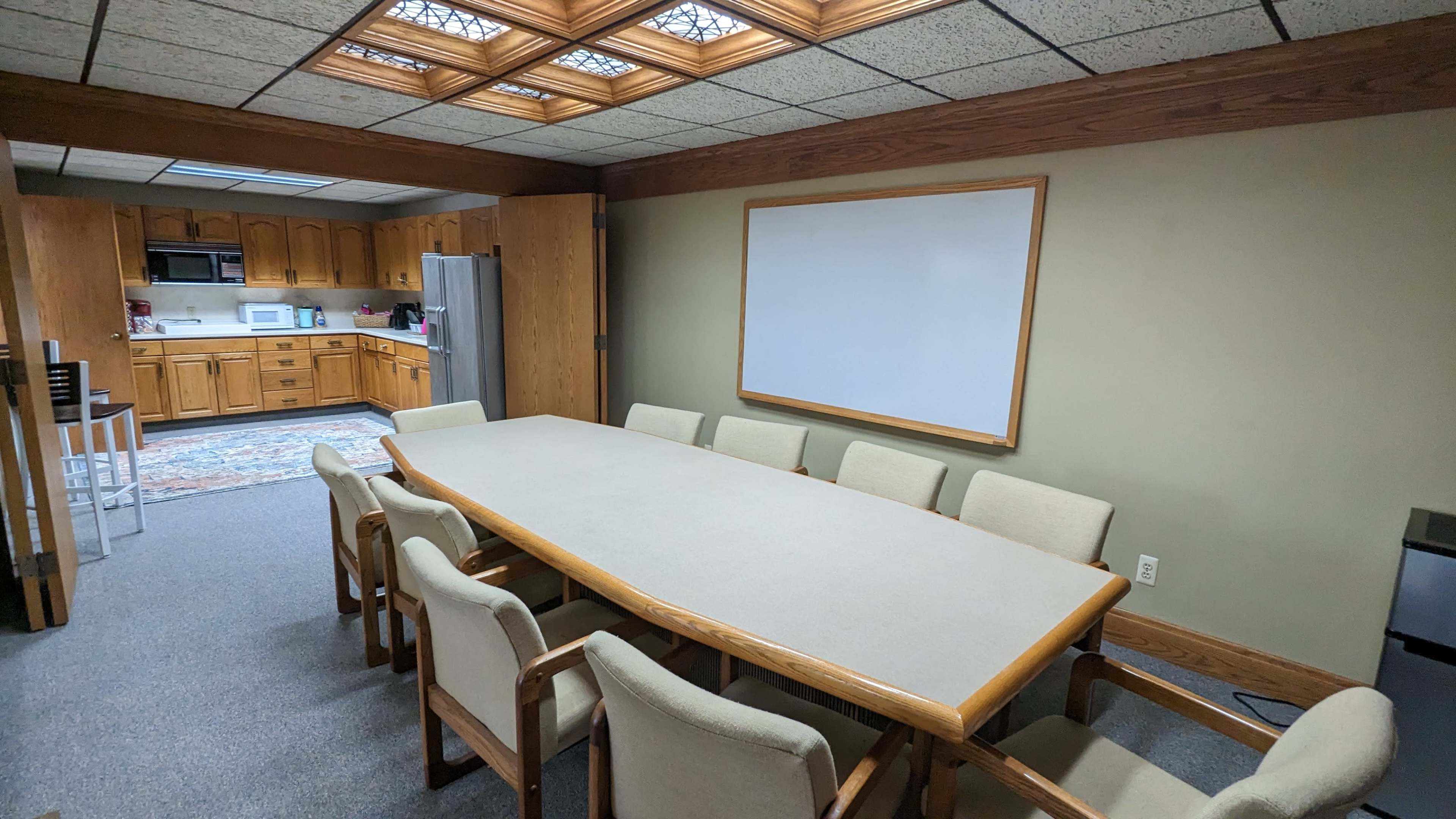 A conference room with a large table surrounded by eight chairs, adjacent to a kitchen area.