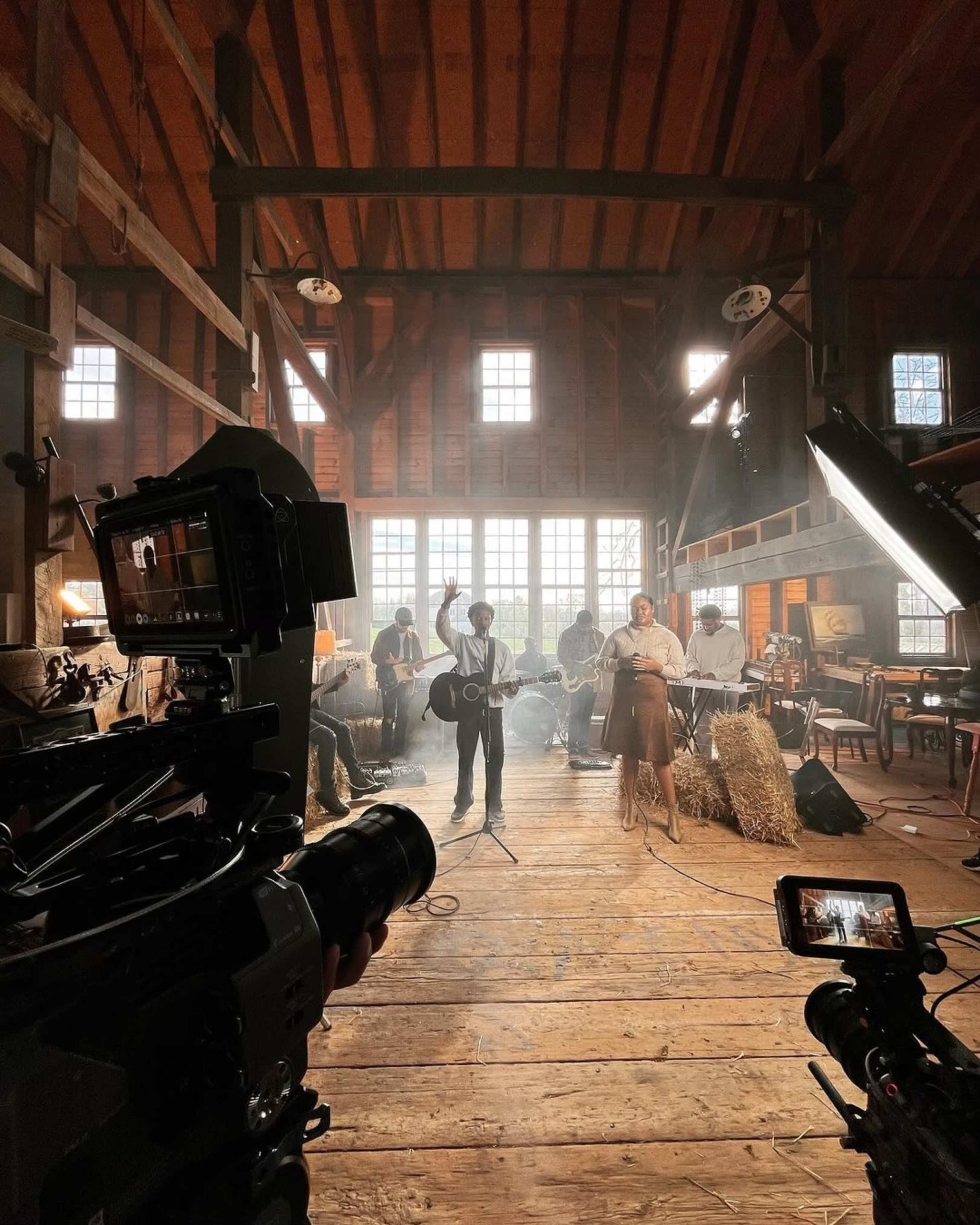 A musician stands in a barn-like space with a band, surrounded by cameras and lights.