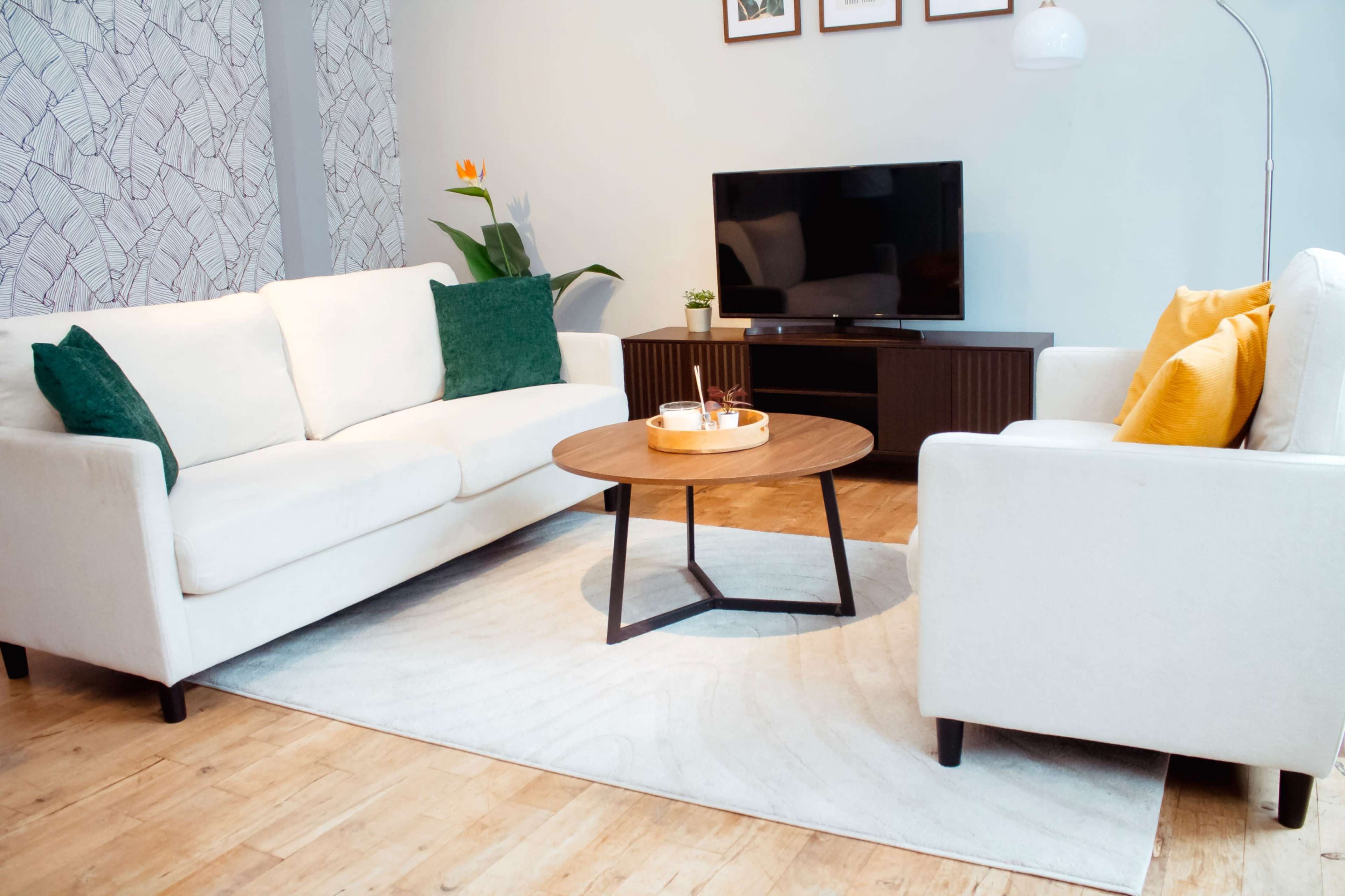 A modern living room features two white sofas with colorful cushions, a wooden coffee table, and a flat-screen TV on a dark-colored media unit.