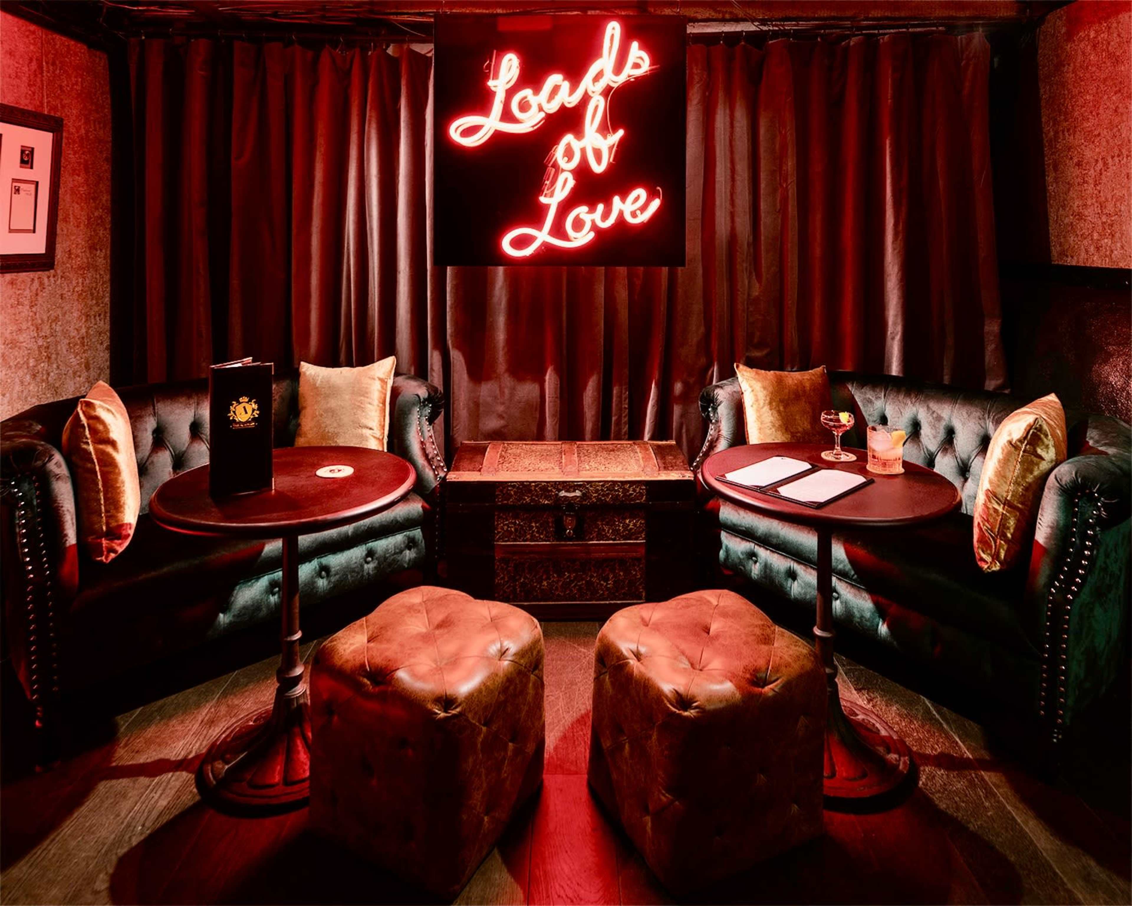 A dimly lit lounge features plush dark green sofas, two small round tables, and a neon sign reading "Loads of Love" against a backdrop of deep red curtains.