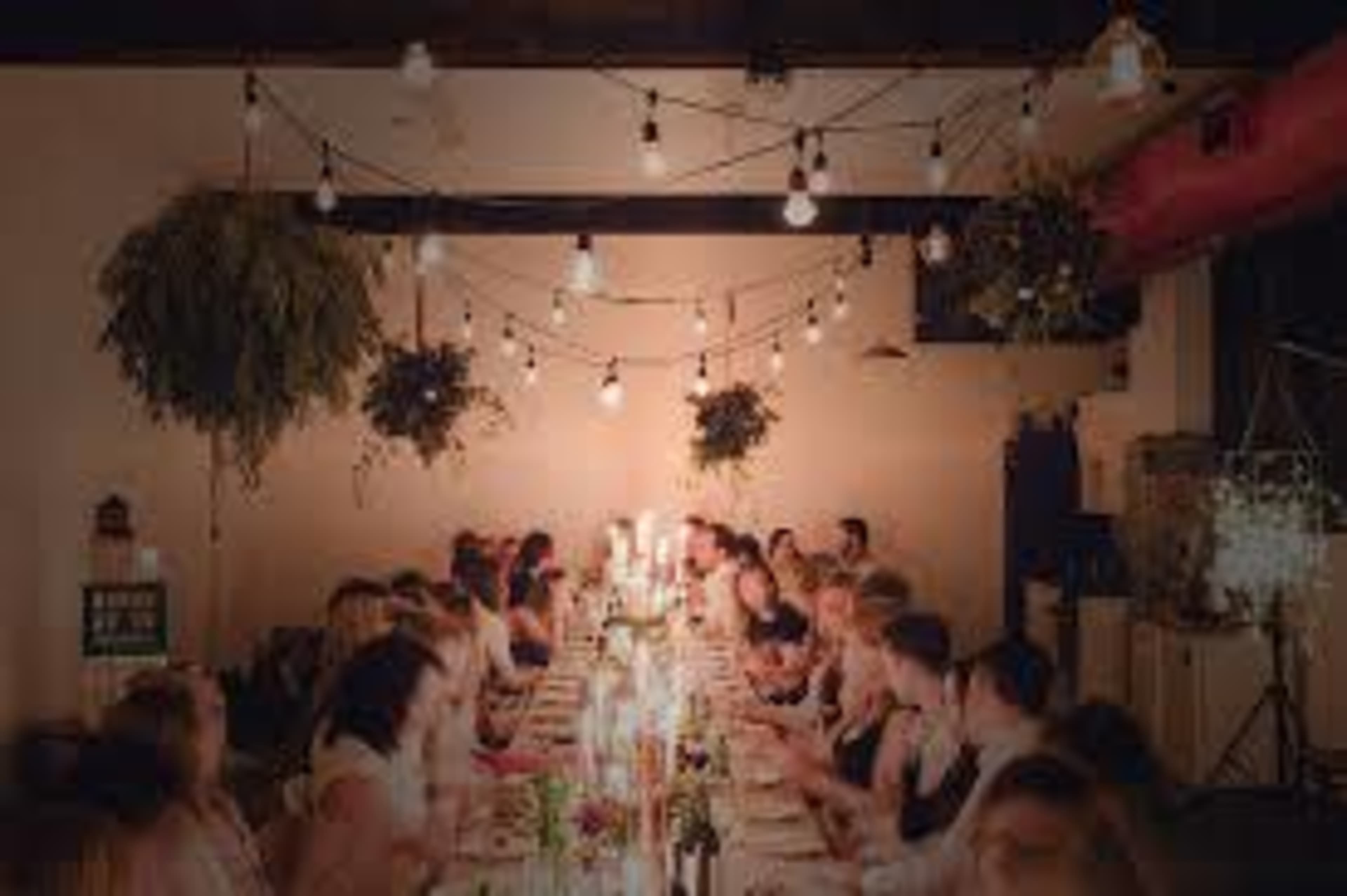 A long table is set for a dinner party, lined with candles and surrounded by guests in a warmly lit space with hanging plants and string lights overhead.