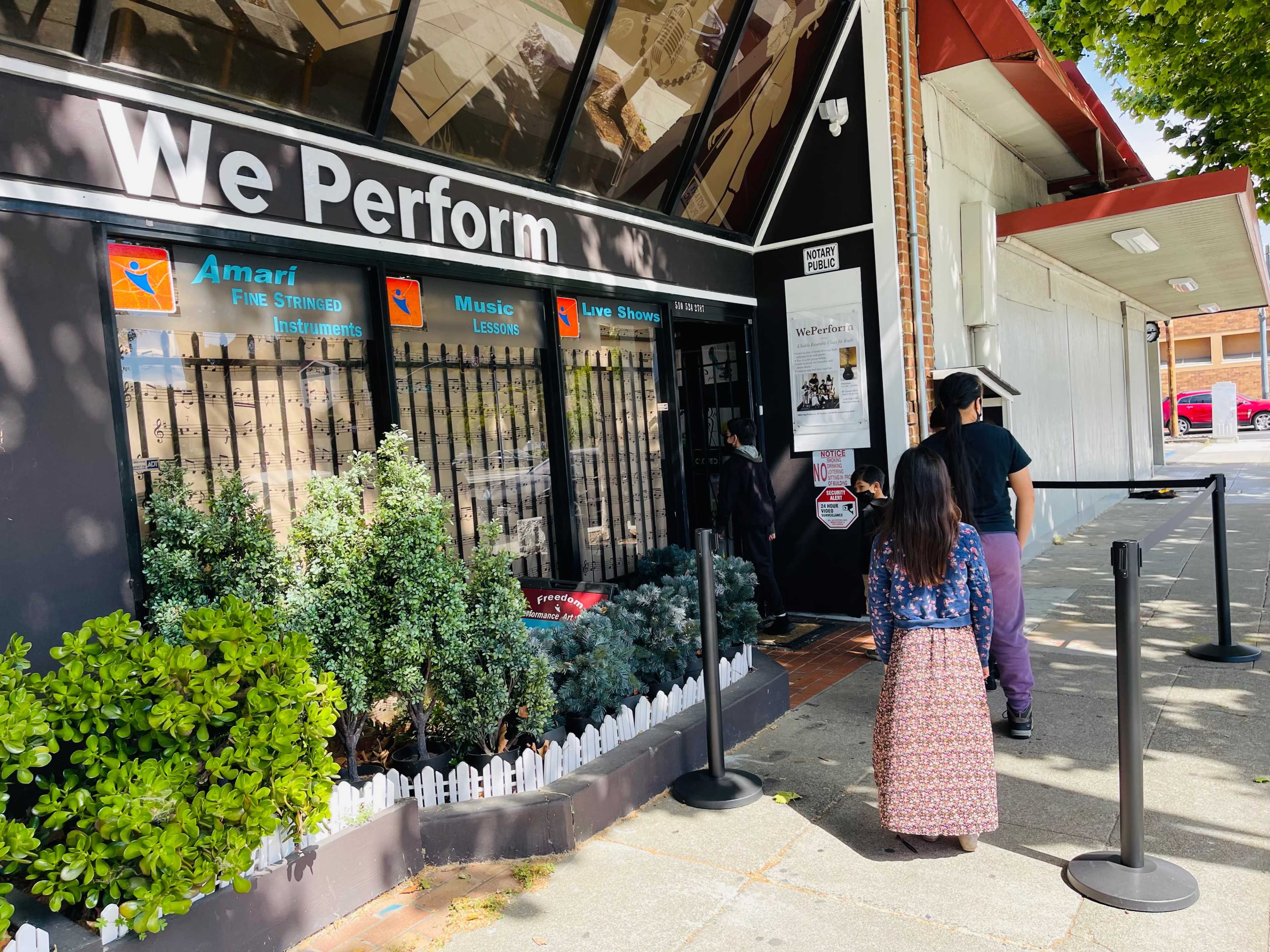 A woman and a child walk toward a storefront labeled "We Perform," which features a large glass window displaying various signs.