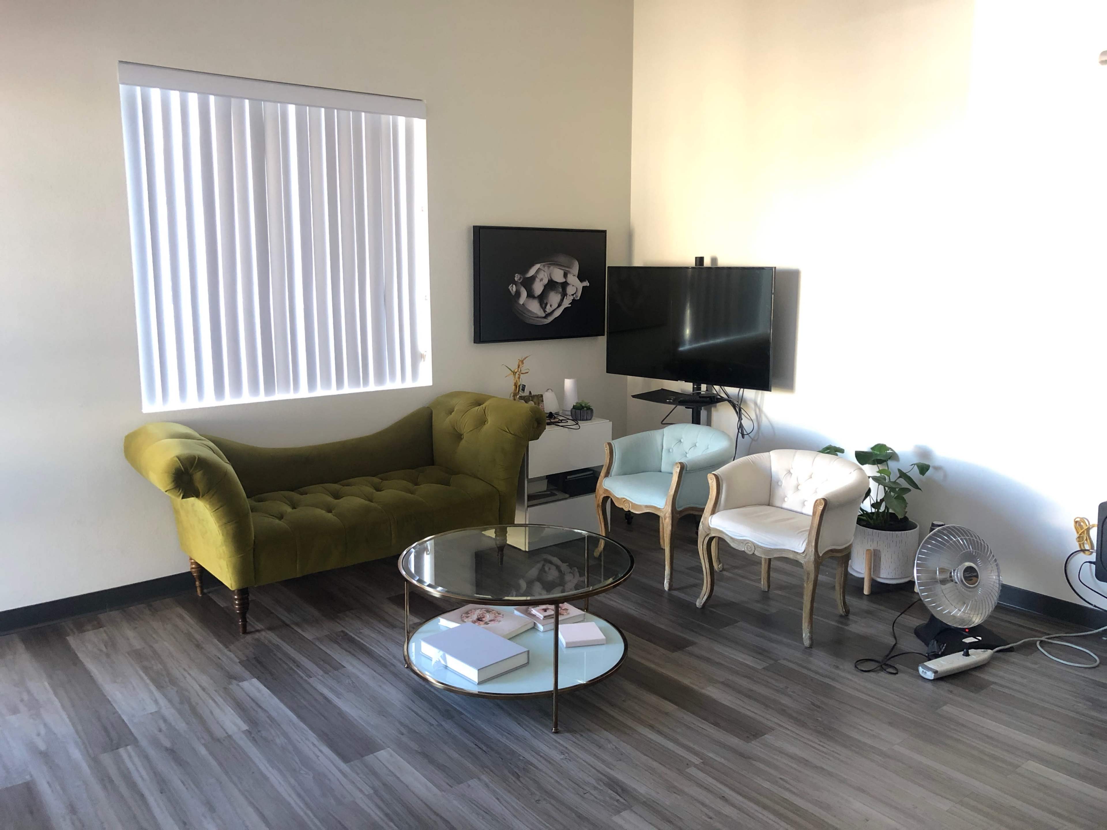 A living room with a green velvet couch, two white chairs, a round coffee table, a television on a stand, and a large window with vertical blinds.