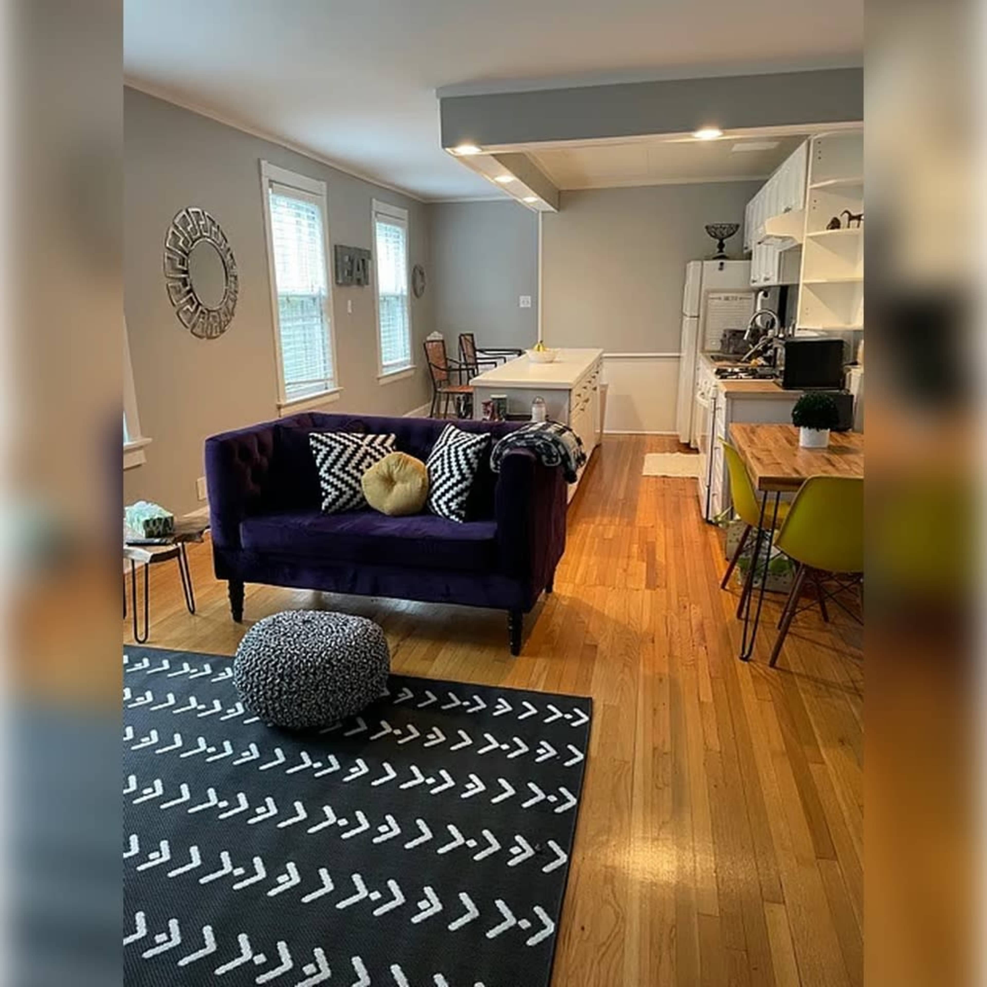 The image shows a living area with a purple couch, a wooden dining table, and a kitchen in the background, all arranged on a patterned area rug.