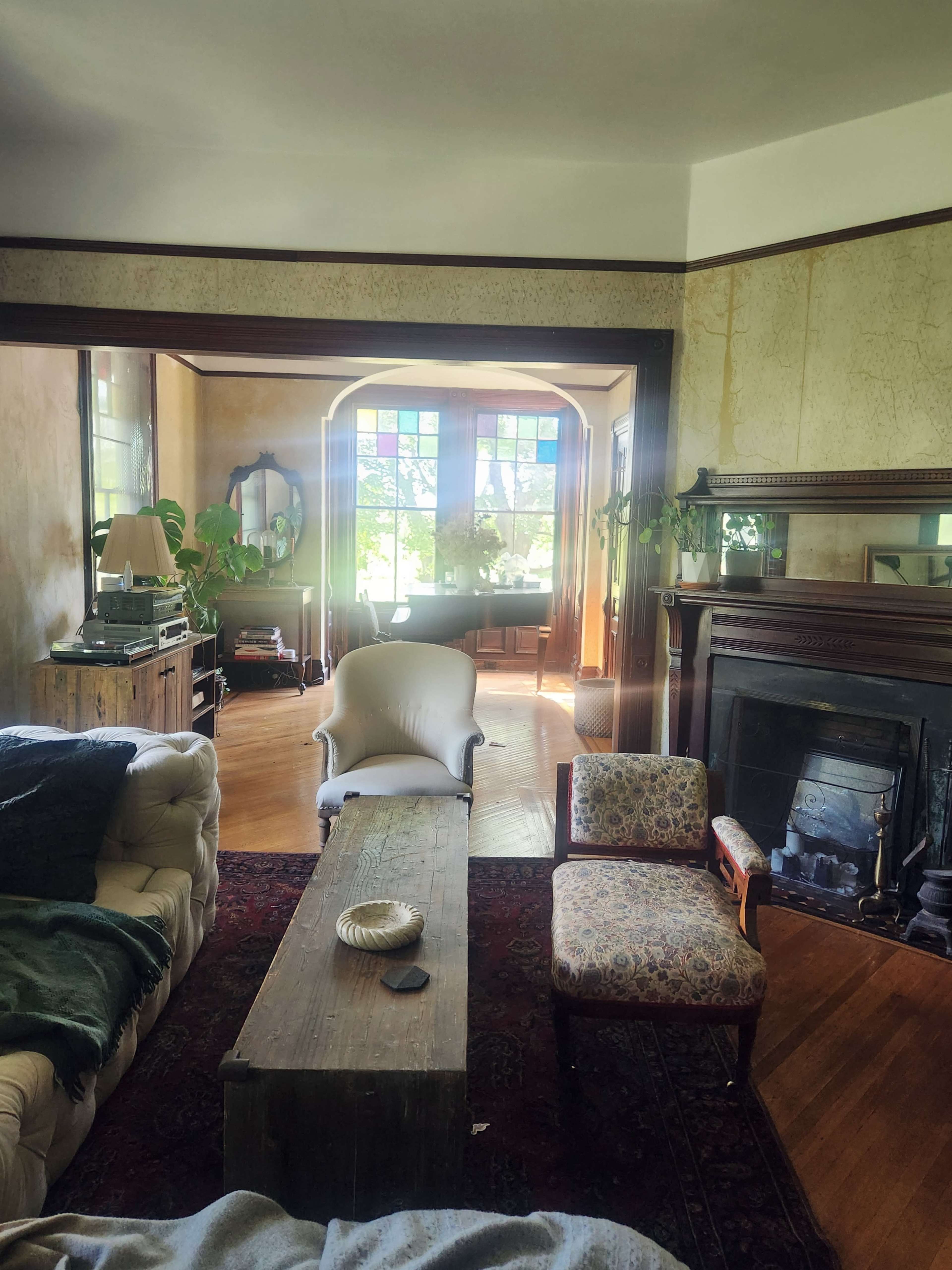 The image shows a cozy living room with a mix of vintage furniture, a large wooden table, and a sunlit area filled with plants.