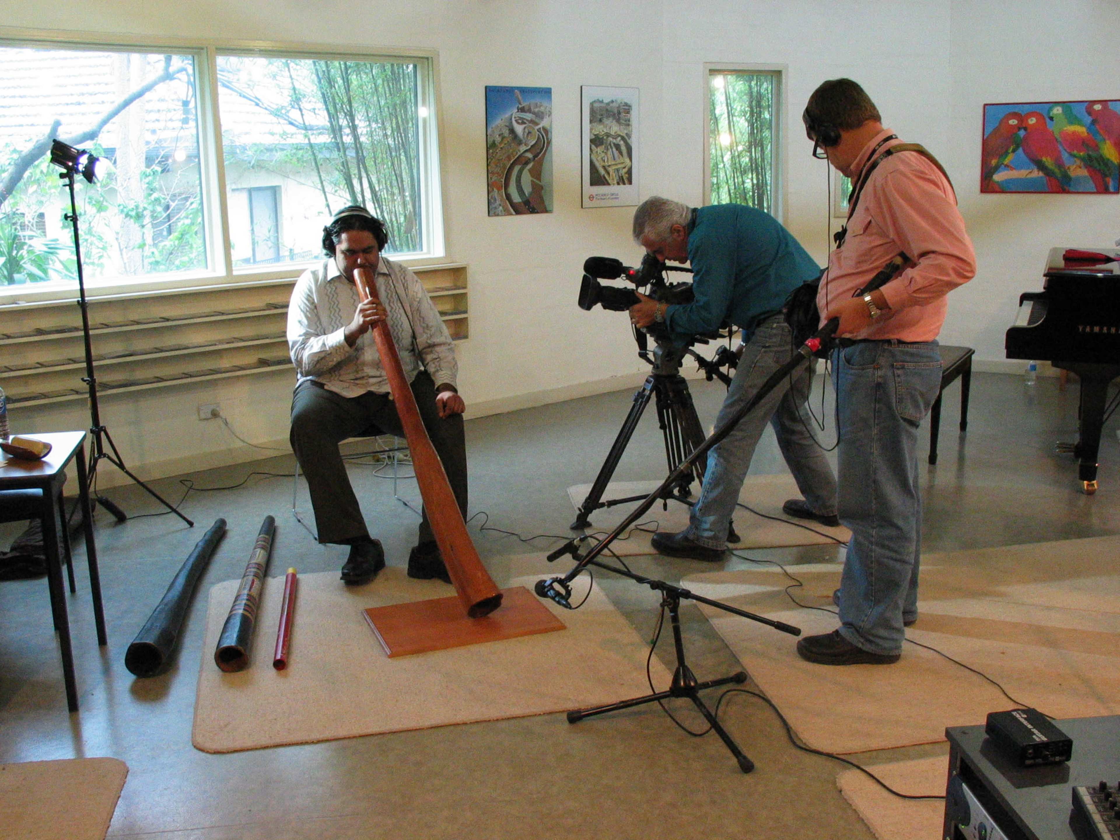 A man plays a didgeridoo while two cameramen record the performance in a room with large windows and colorful artwork.