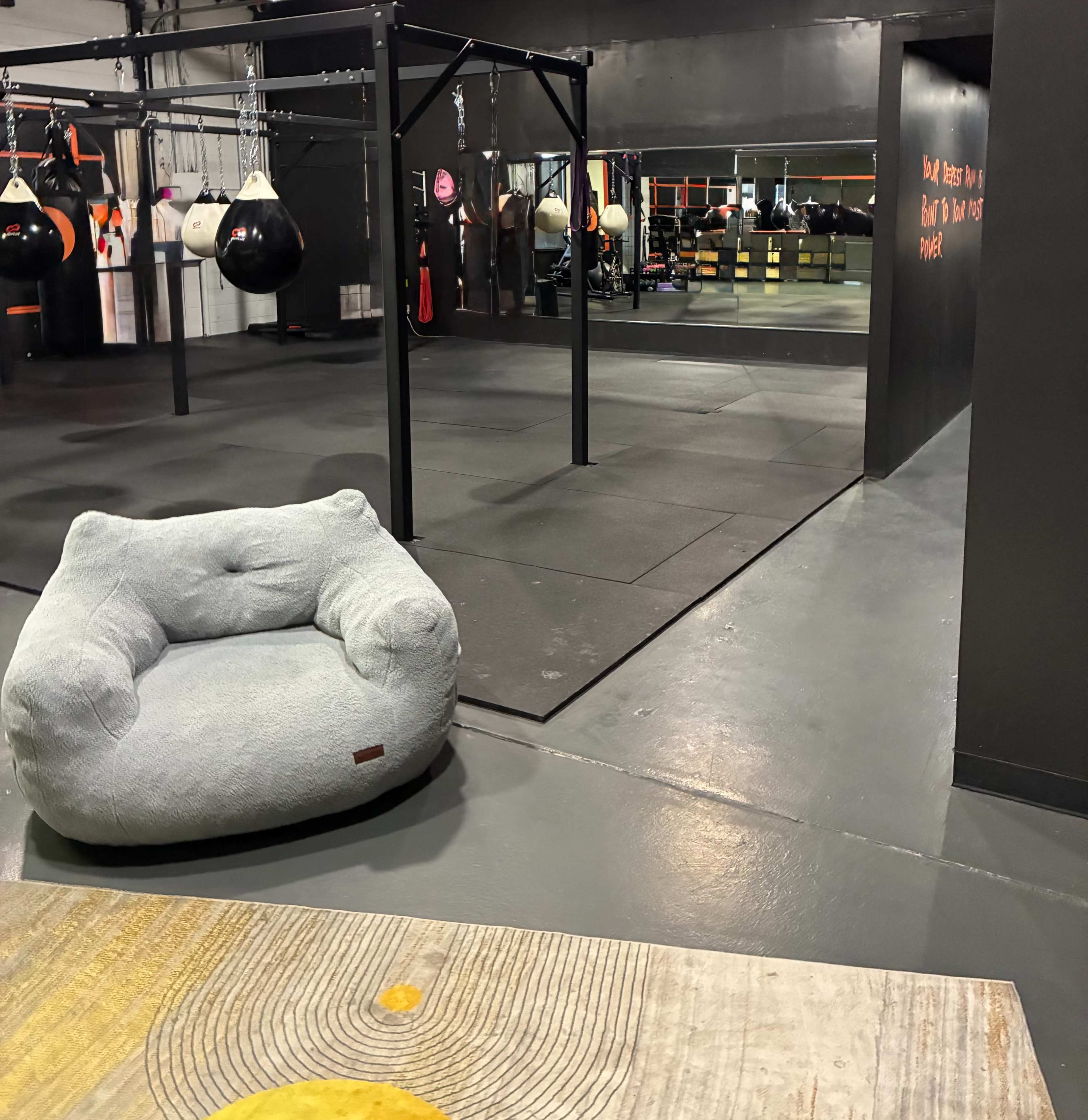 The image shows a gym interior featuring boxing bags, exercise equipment, and a grey bean bag chair positioned near the entrance.