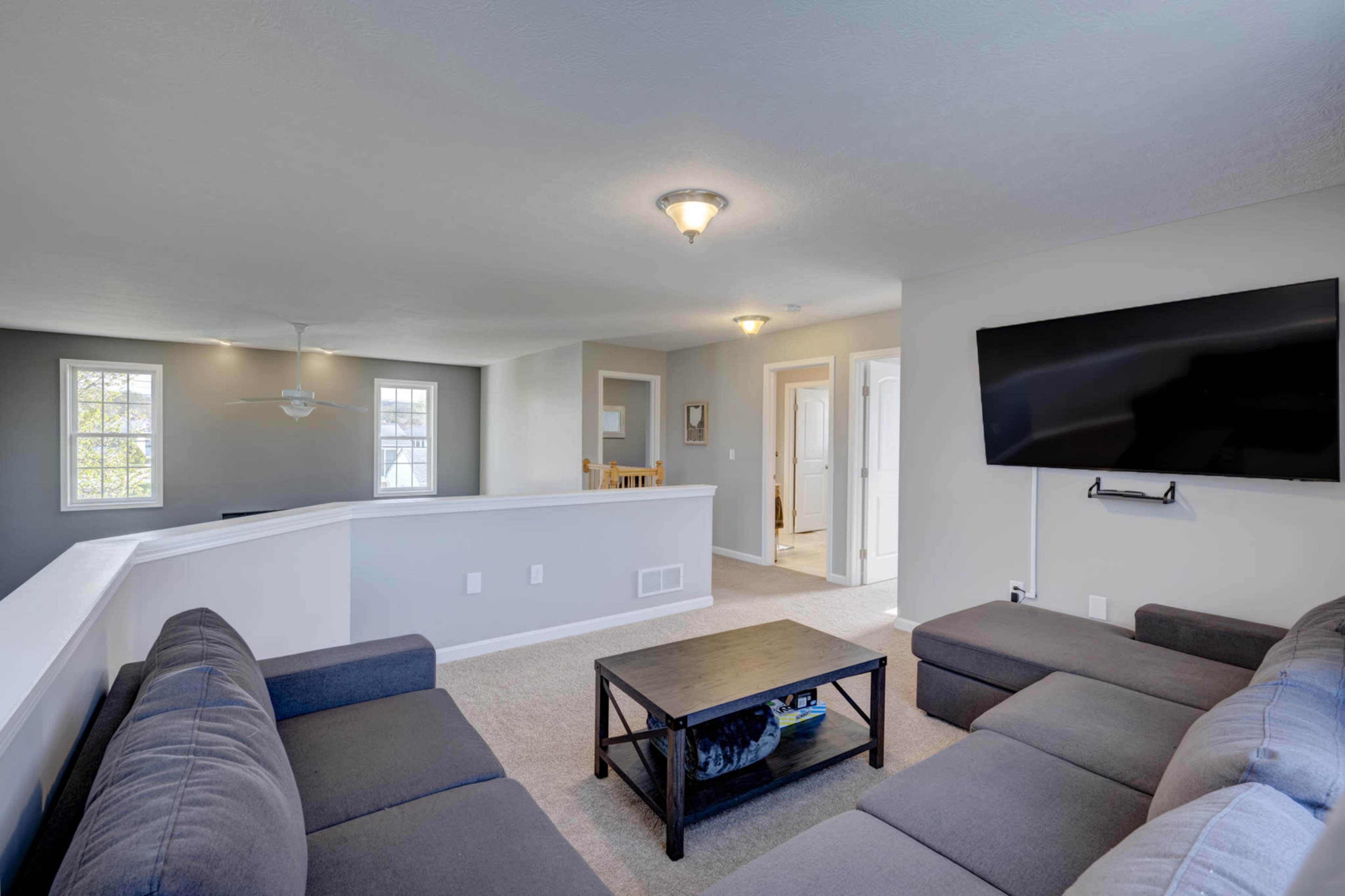 A modern living room with a sectional couch, a coffee table, and a wall-mounted television, connected to an open space with light-colored walls and windows.