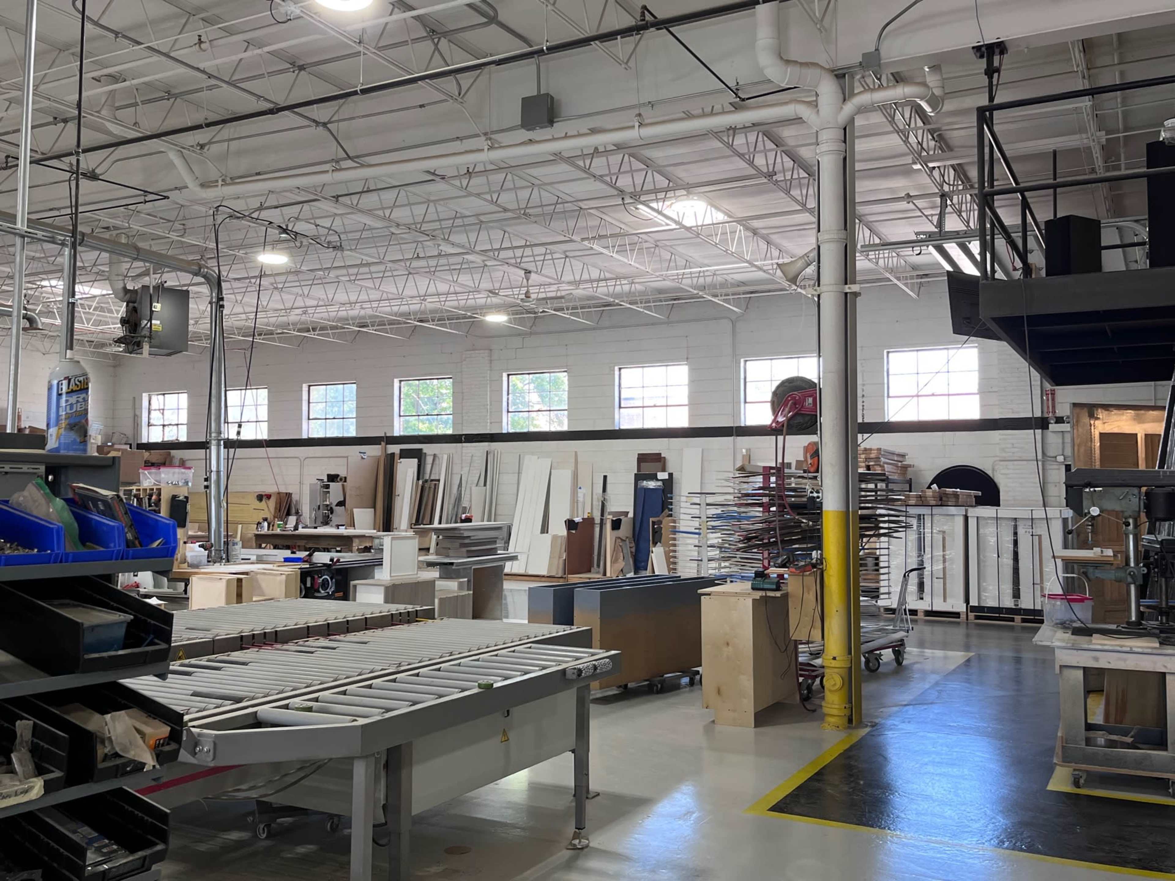 A spacious workshop with various woodworking materials, tools, and machinery organized throughout the area.