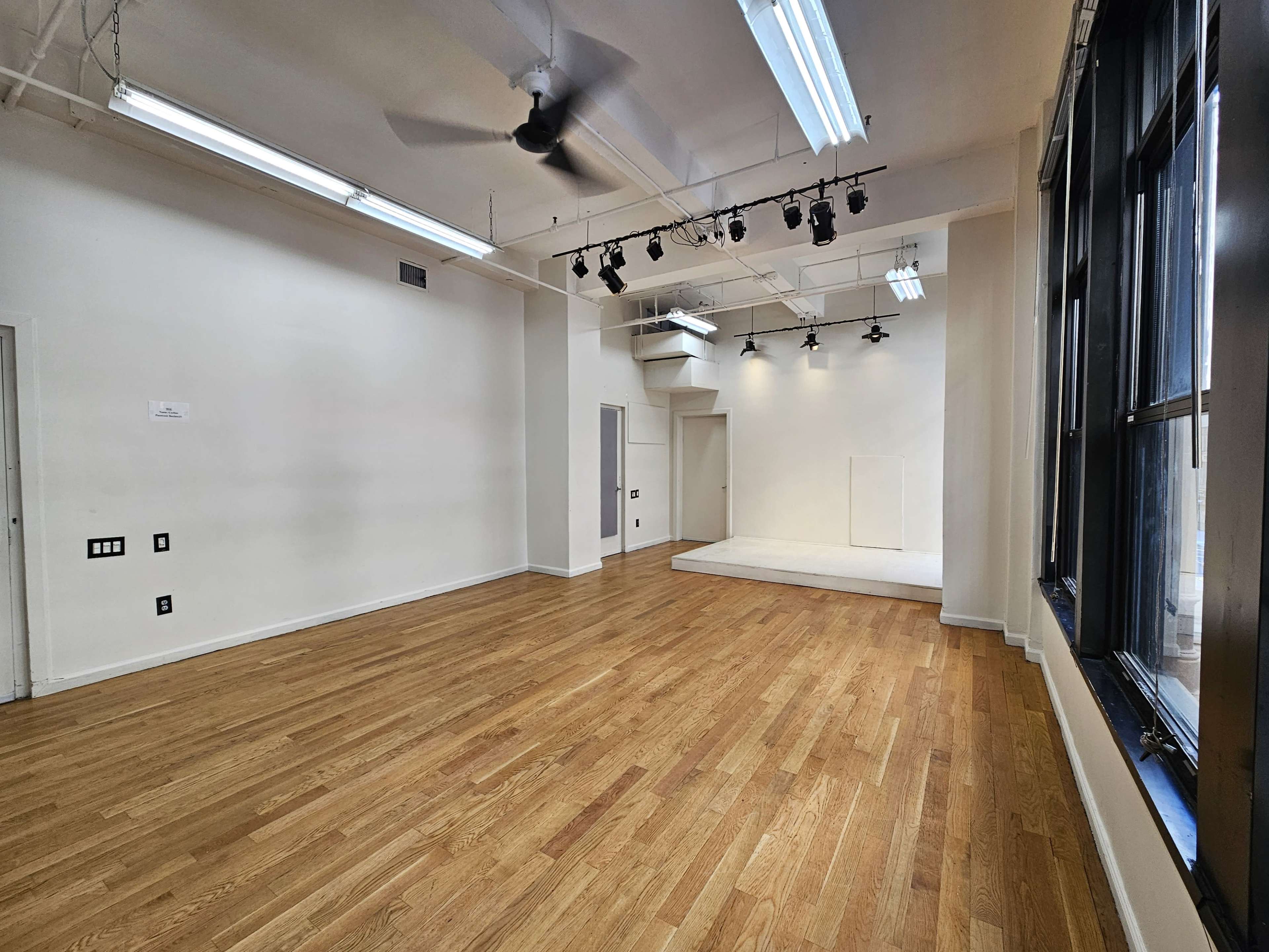 An empty room with hardwood floors and large windows, featuring ceiling lights and a small elevated platform at one end.