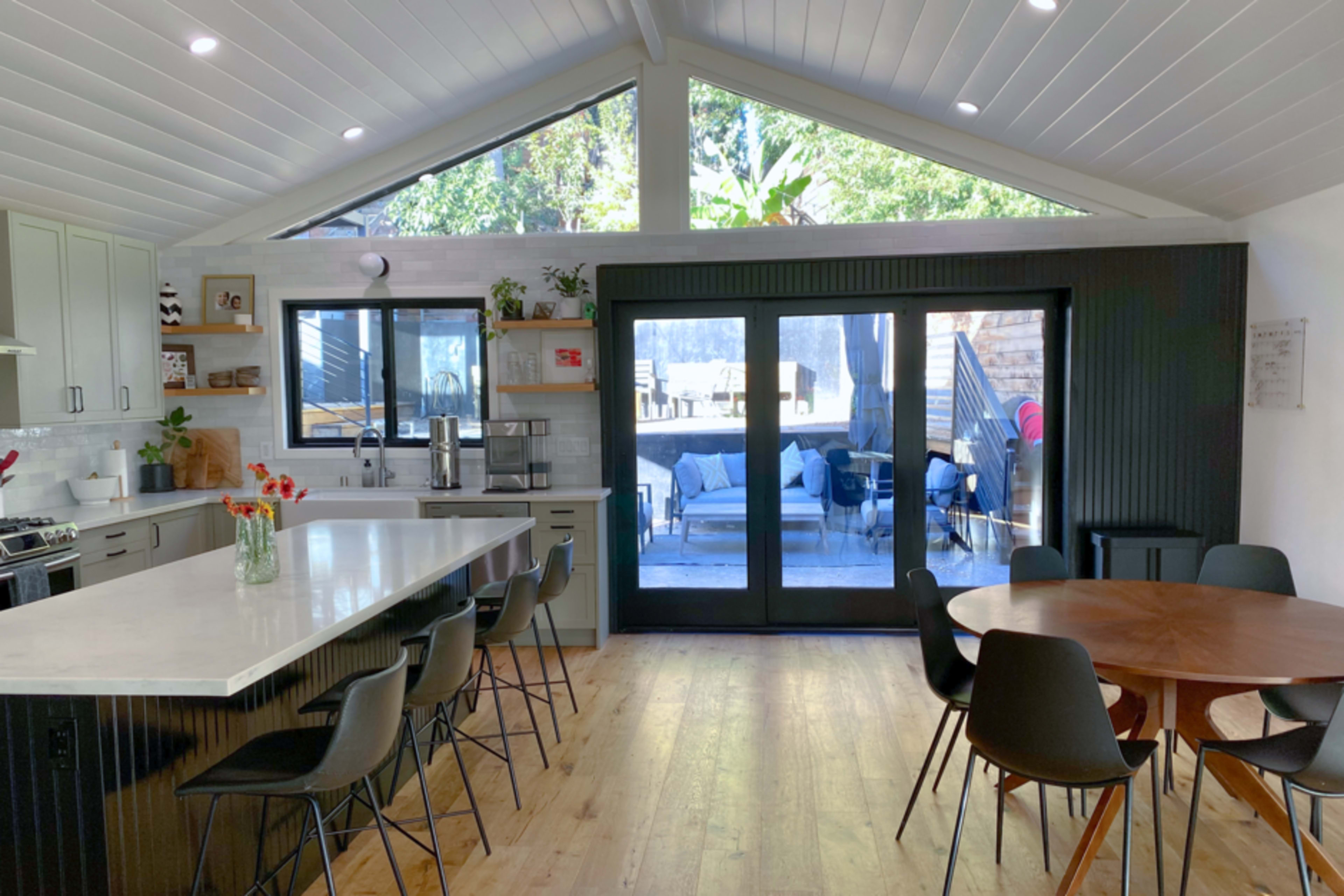 A modern kitchen features a large island with seating, a dining table, and large windows that open to an outdoor seating area.