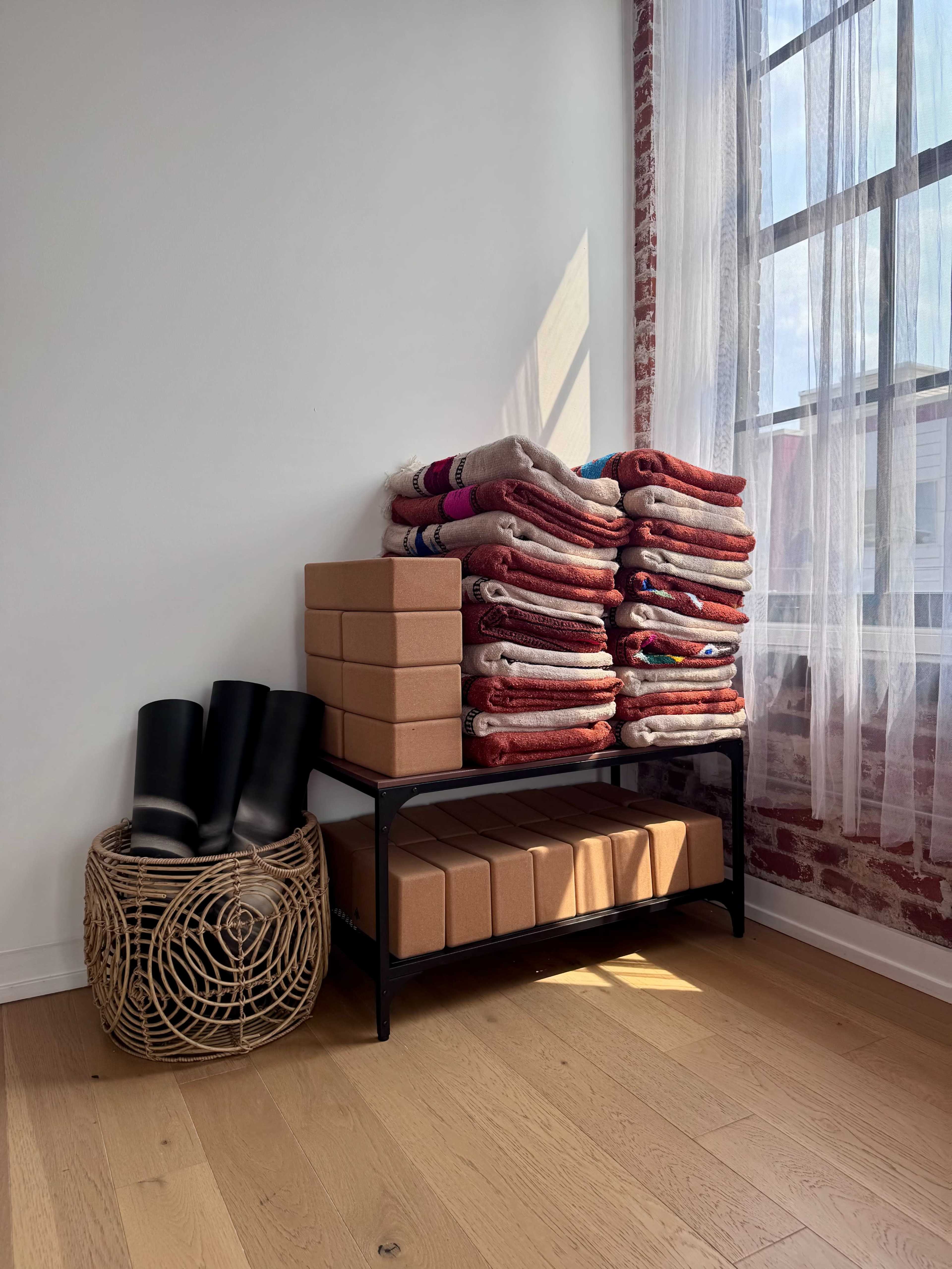 A stack of neatly arranged blankets and yoga blocks is placed on a black metal shelf beside a woven basket containing rubber boots, with sunlight streaming through a large window.