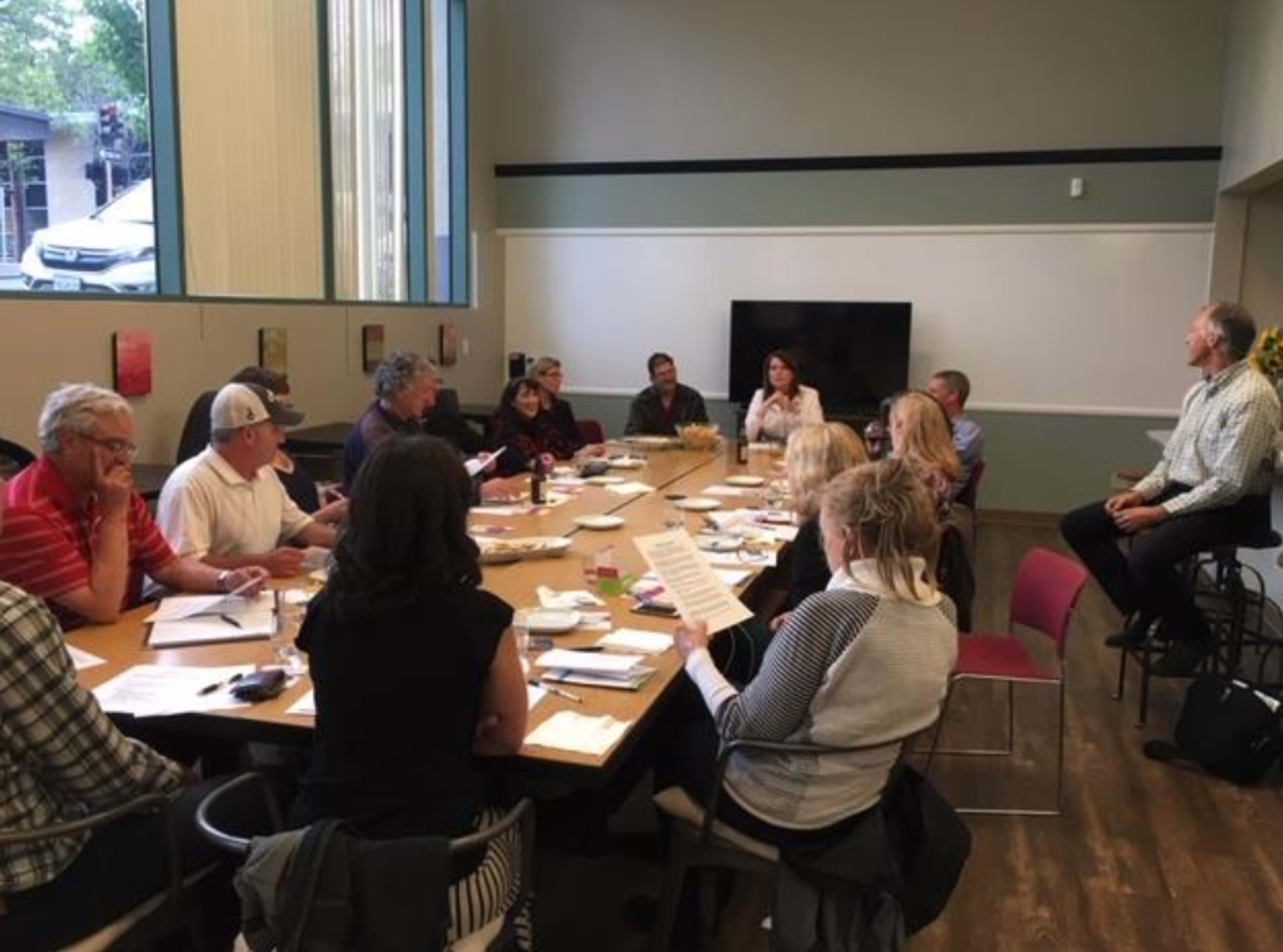 A group of approximately fifteen people is seated around a large conference table, engaged in a meeting with papers and materials in front of them.