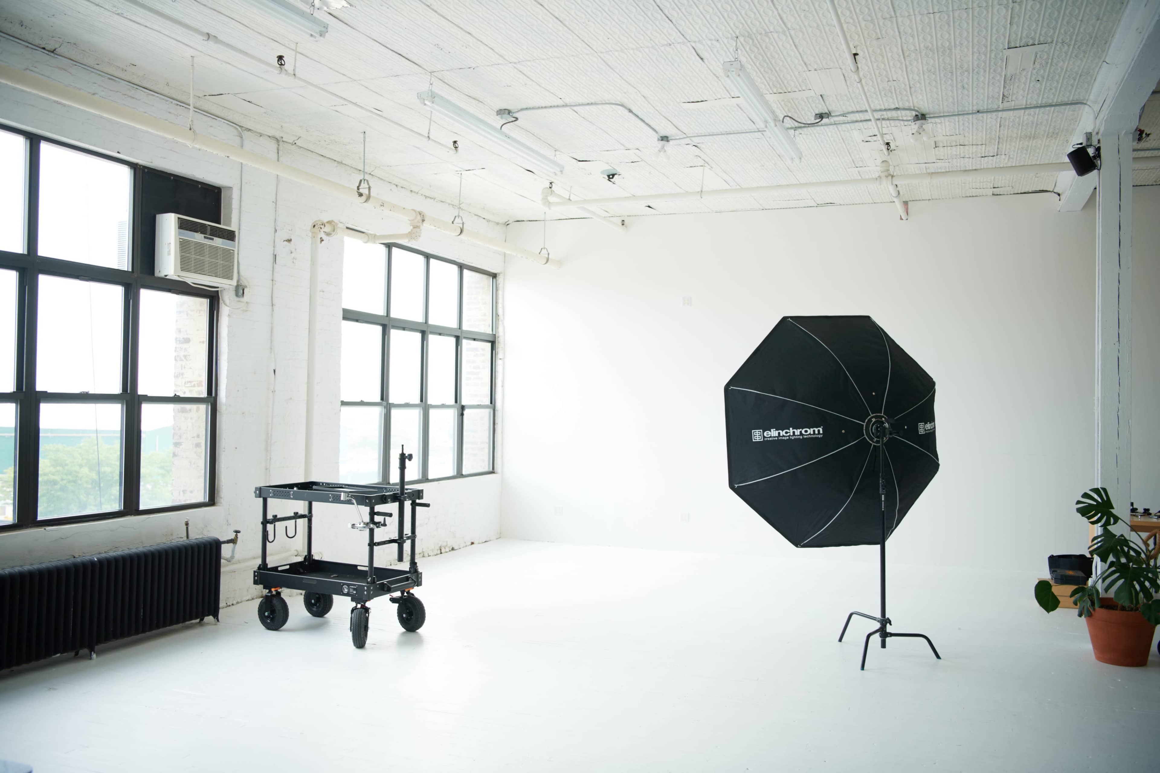 The image shows a spacious, well-lit studio with large windows, an octagonal softbox light on a stand, and a wheeled cart in the corner.