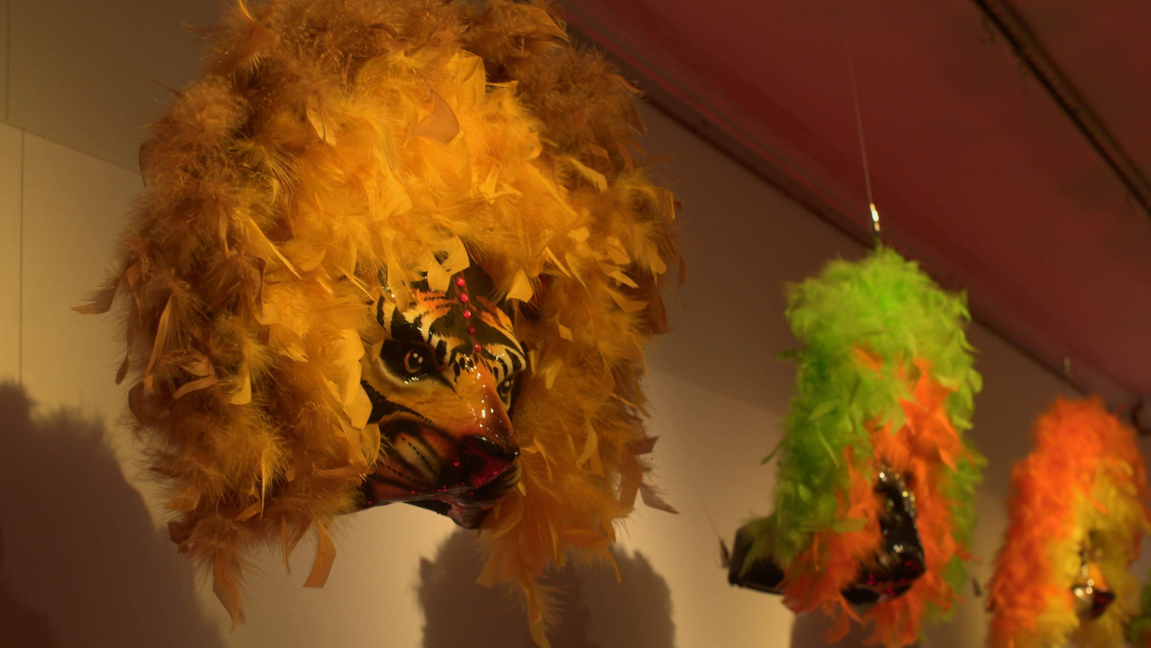 Two theatrical masks adorned with vibrant feathers, one in yellow resembling a lion and the other in green, hang on a wall.