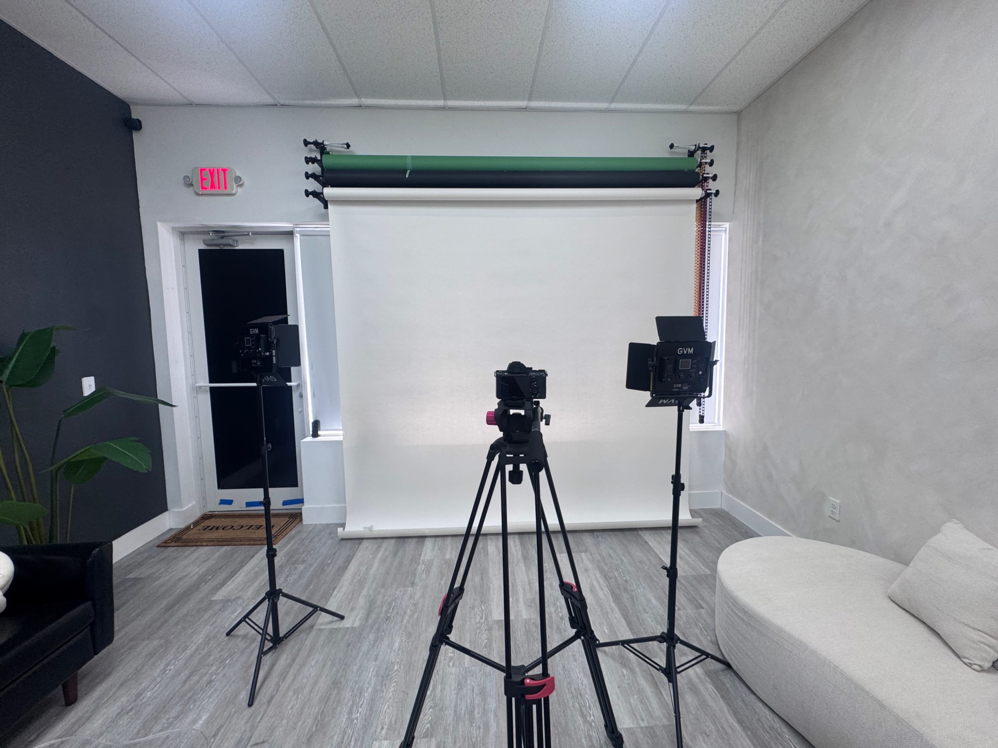 The image shows a photography studio setup with three cameras positioned on tripods facing a white backdrop, surrounded by a chair and plants.