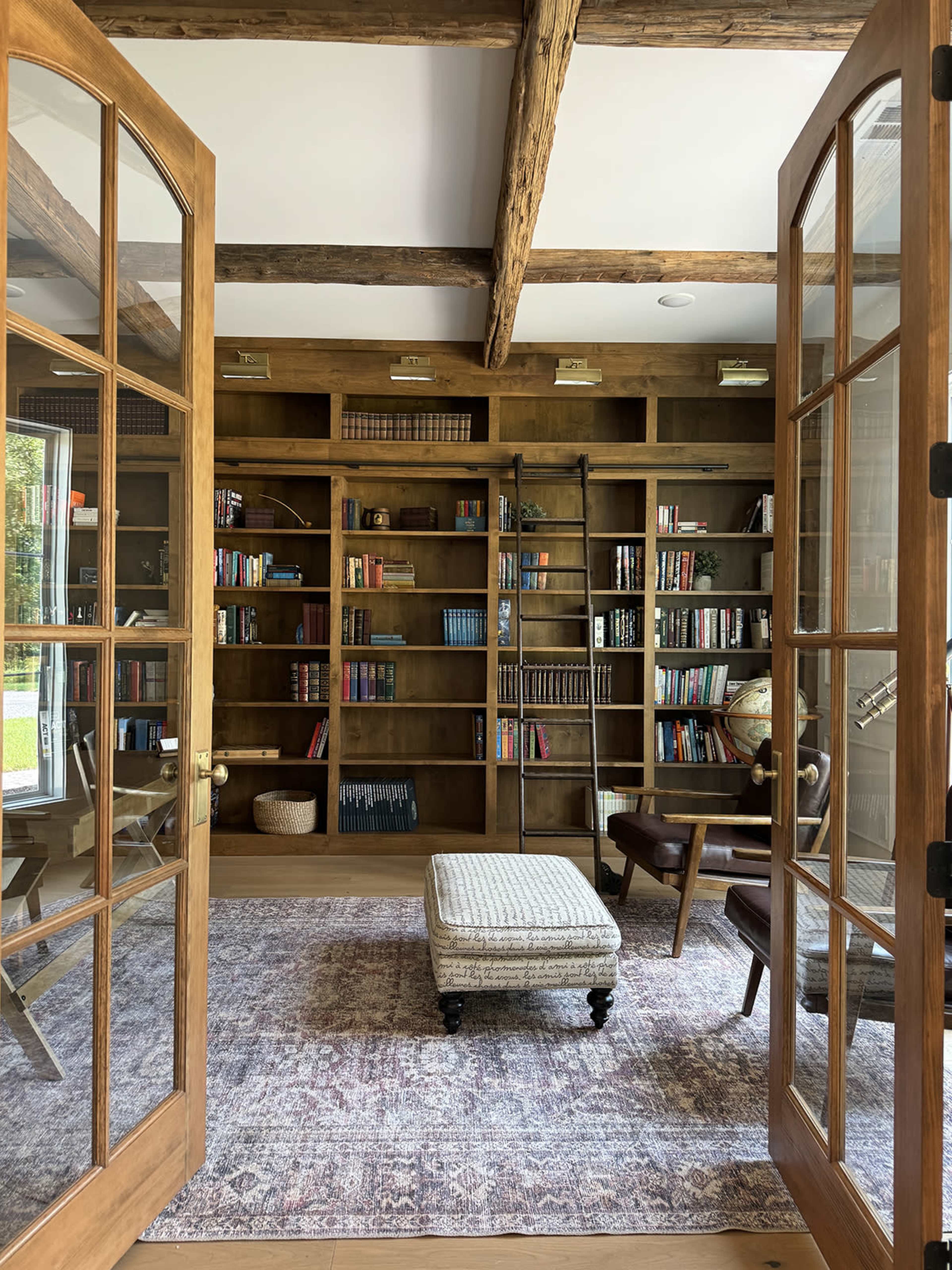 A cozy reading room features wooden shelves filled with books, a tufted ottoman, and a comfortable armchair, all framed by French doors.