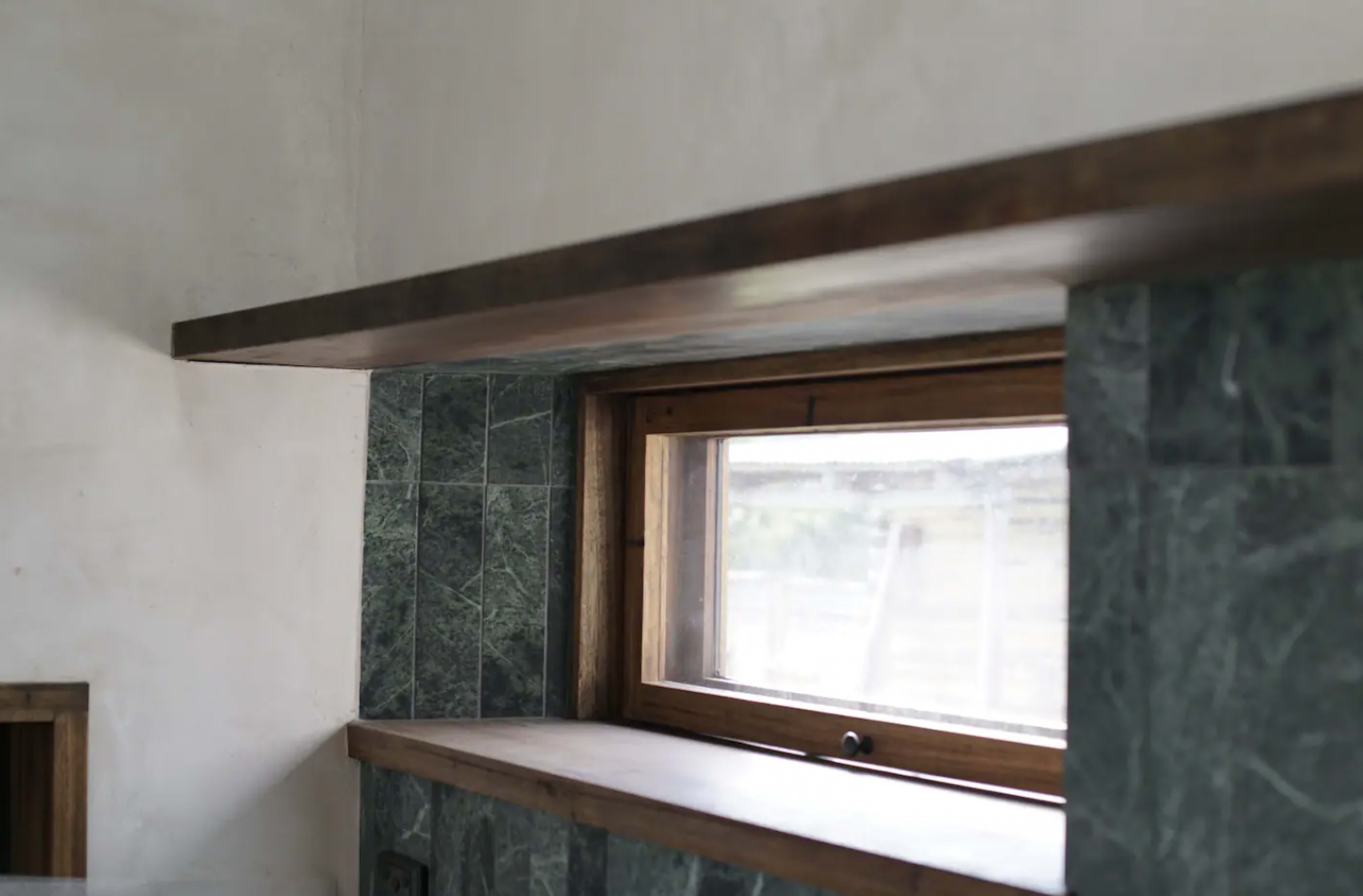 A wooden shelf is installed above a green stone countertop near a window.