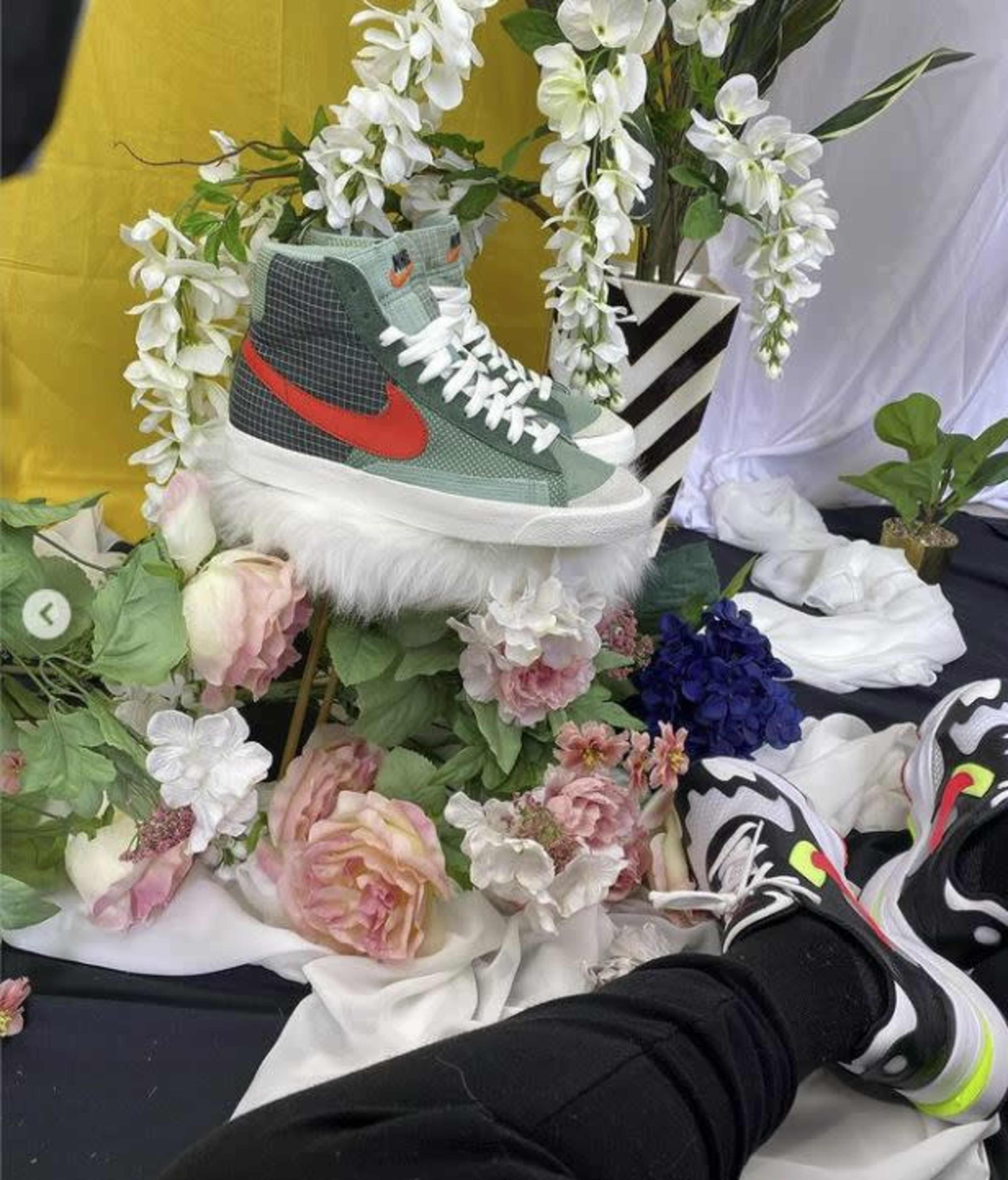 A pair of stylized sneakers sits on a fluffy pedestal surrounded by vibrant flowers and greenery against a backdrop of fabric drapes.