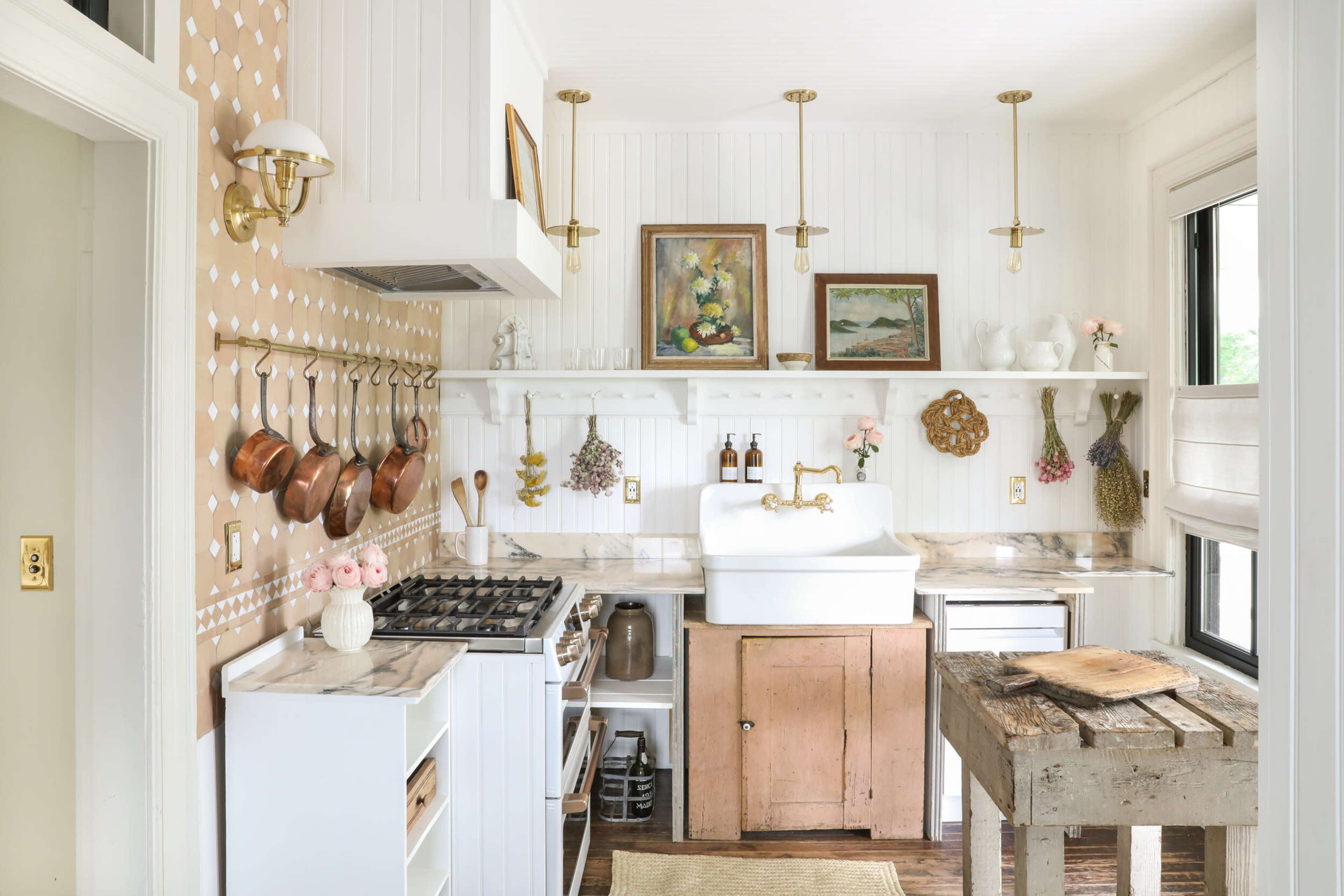 A bright kitchen features white walls, marble countertops, a farmhouse sink, and wooden accents with hanging pots and decorative artwork.