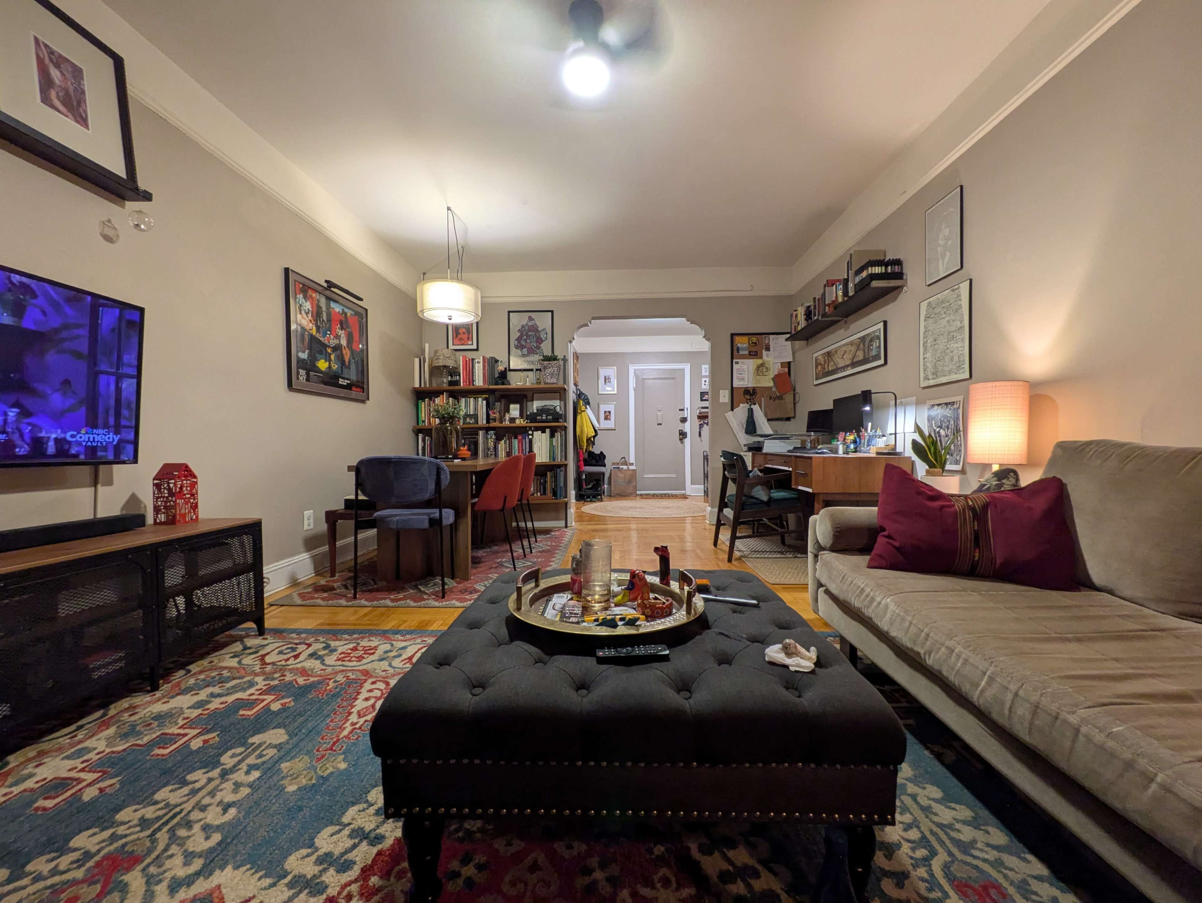 The image shows a cozy living room with a beige sofa, a dark ottoman at the center, a television on the left, and a desk area in the background.