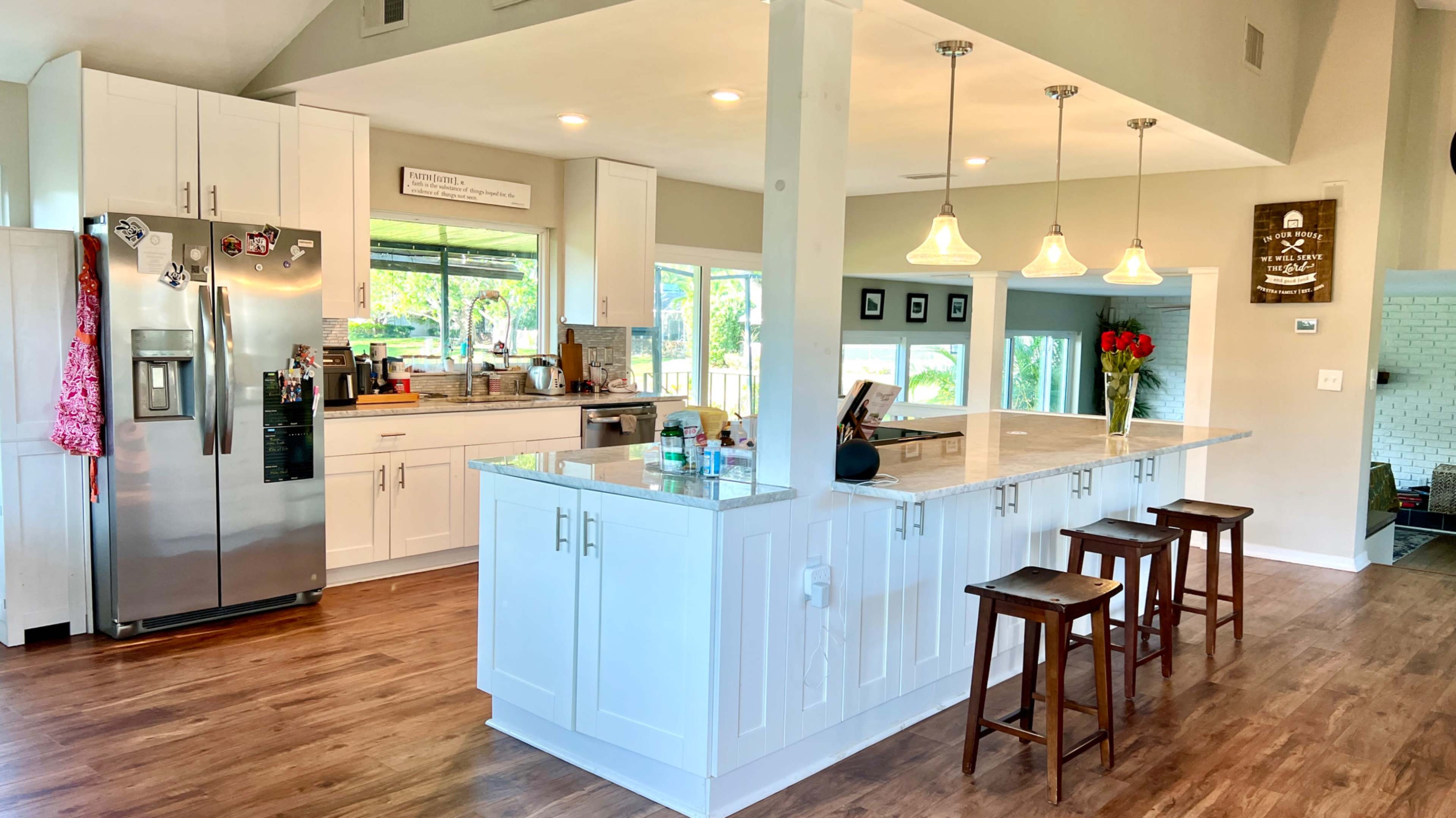 The image shows a modern kitchen with white cabinets, a central island with three wooden stools, and large windows letting in natural light.