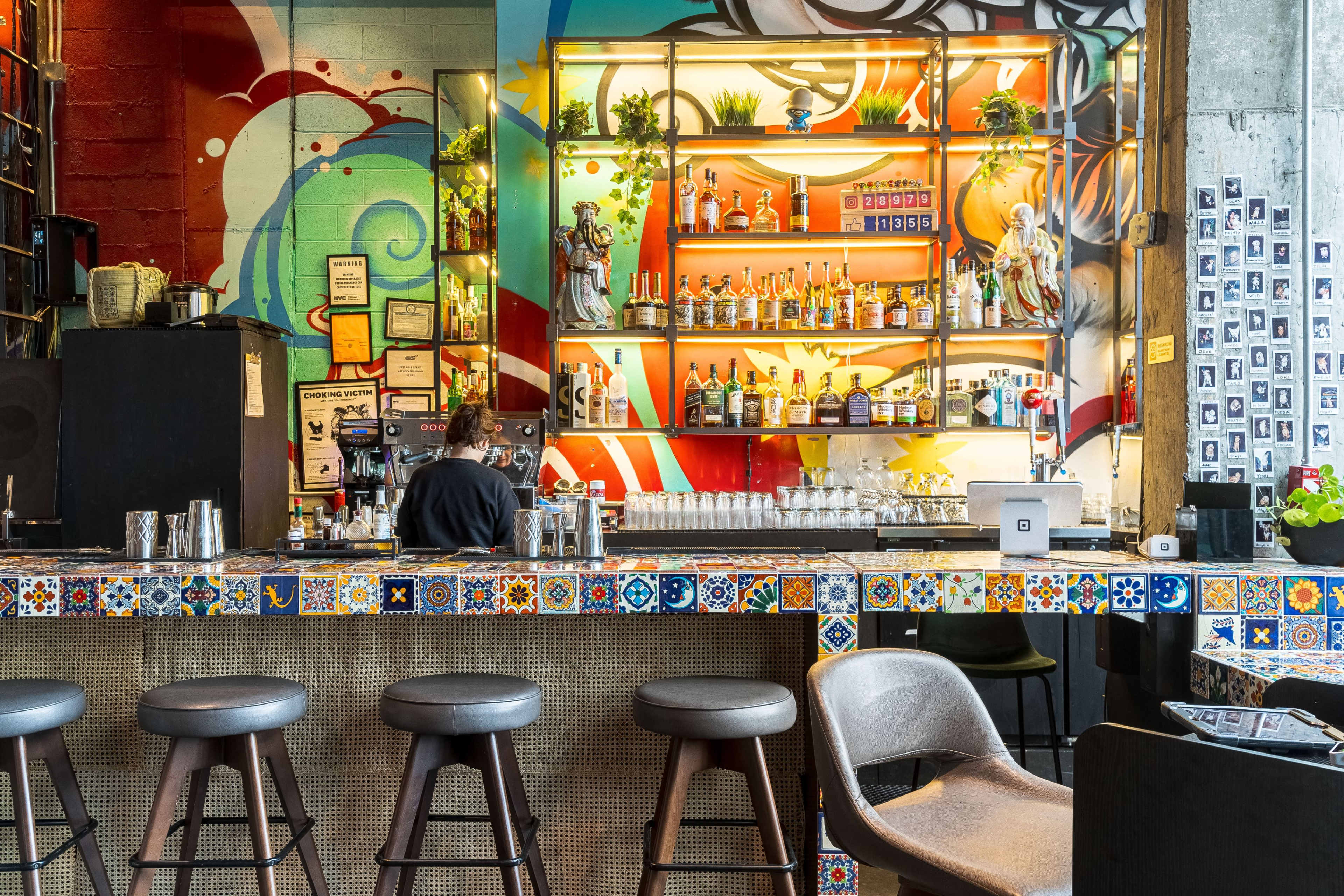 A bartender prepares drinks at a colorful bar adorned with decorative tiles and a vibrant mural in the background.