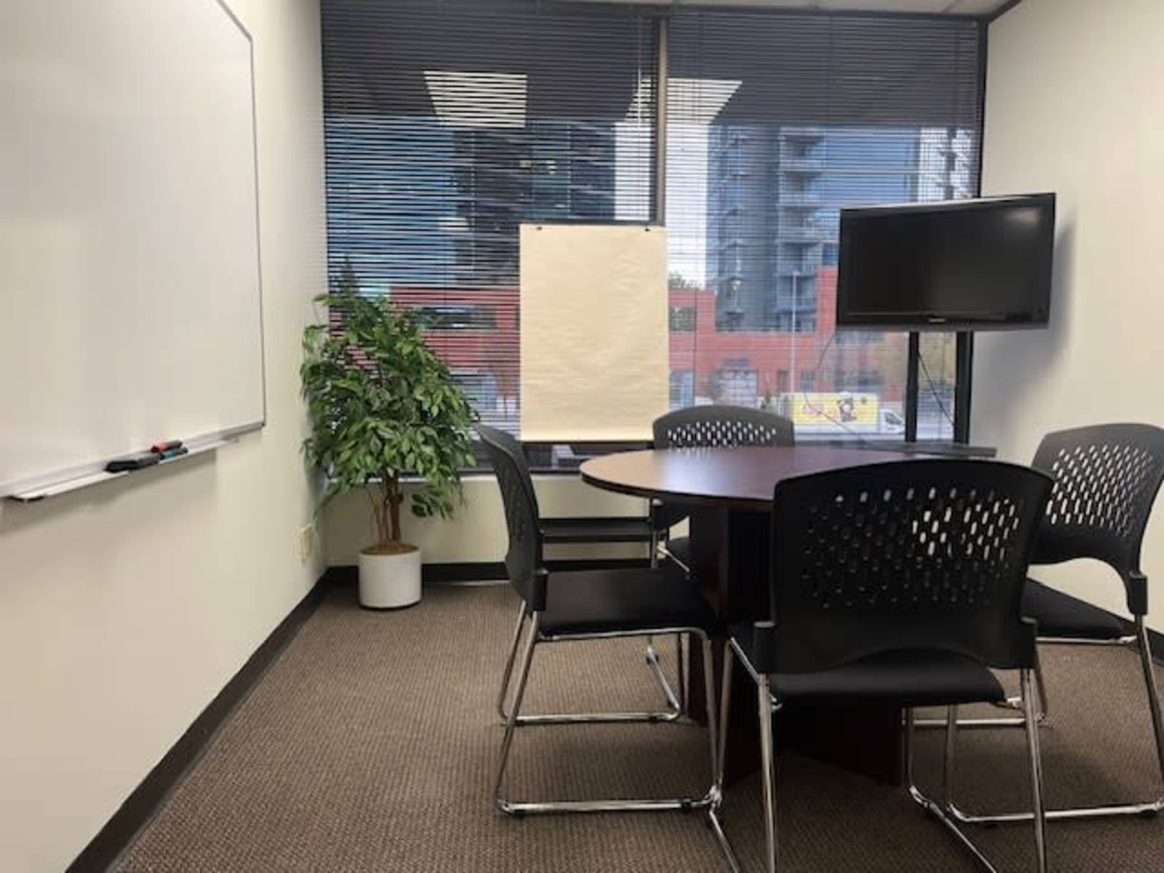 The image shows a conference room with a round table surrounded by black chairs, a whiteboard on one wall, a large monitor on another, and a potted plant near a window.