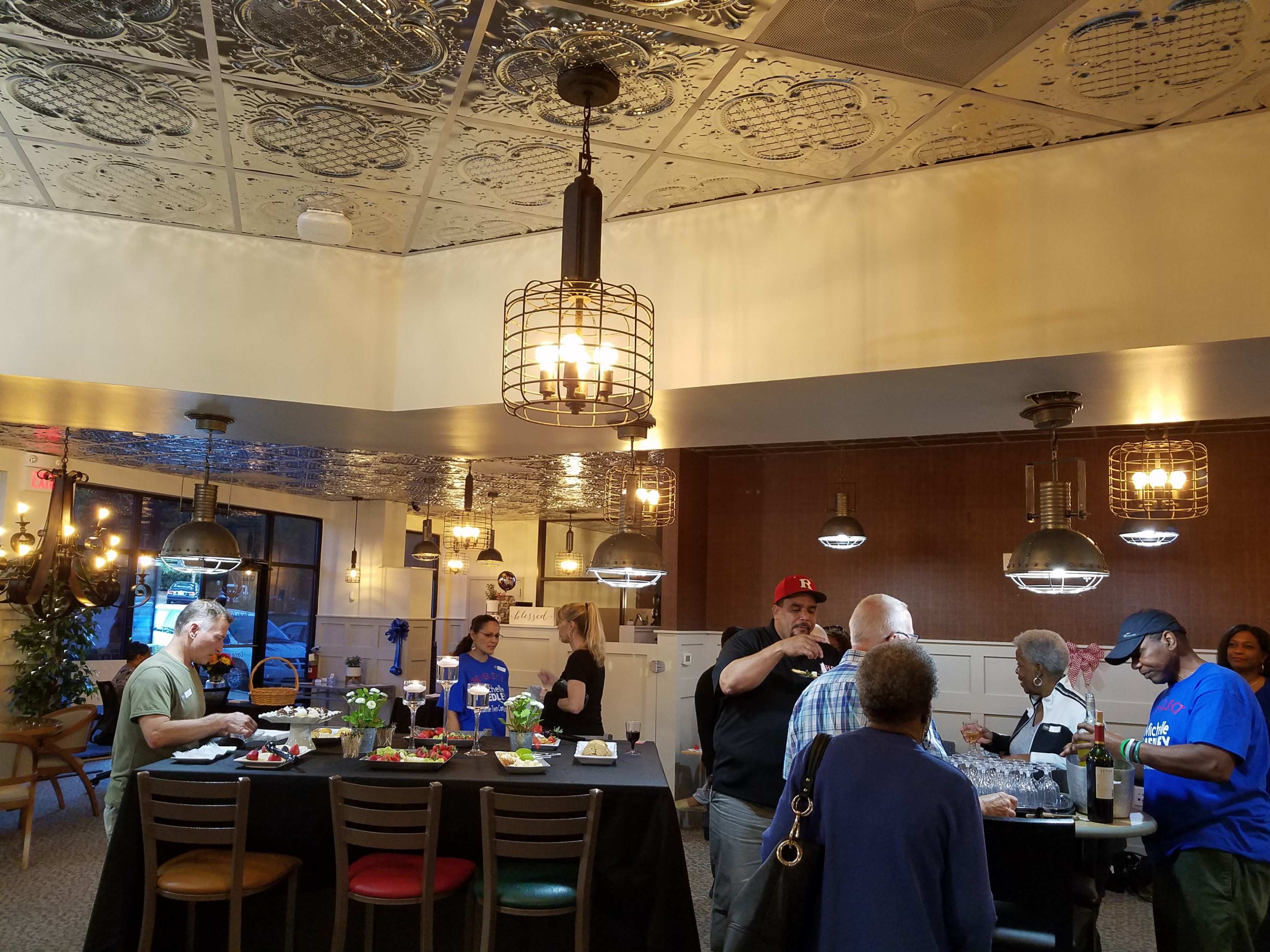 A group of people gathers in a well-lit restaurant with a central table adorned with food and drinks, while others engage in conversation.