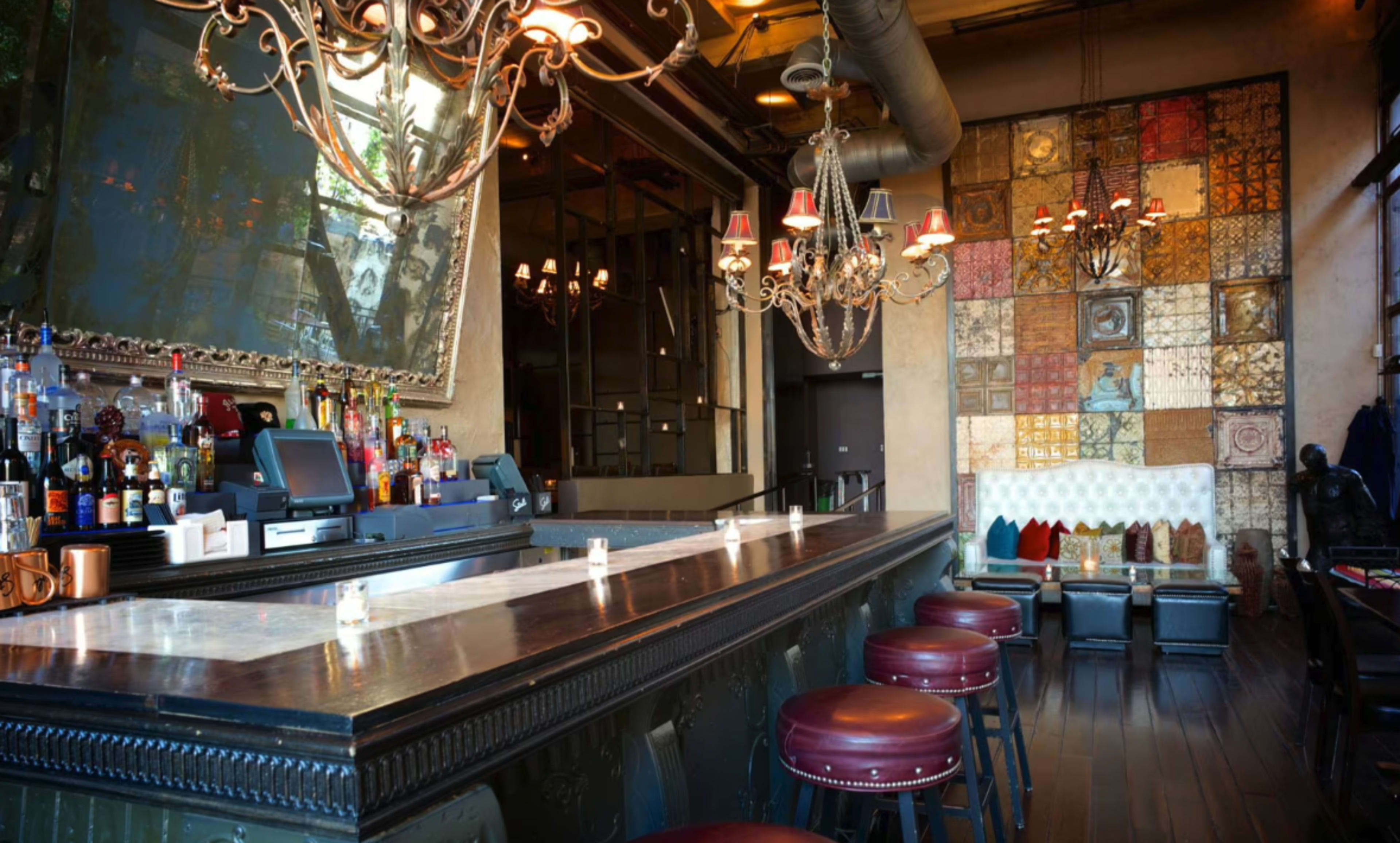 The image shows a stylish bar with a long counter, ornate chandeliers, and a decorative wall featuring colored tiles.
