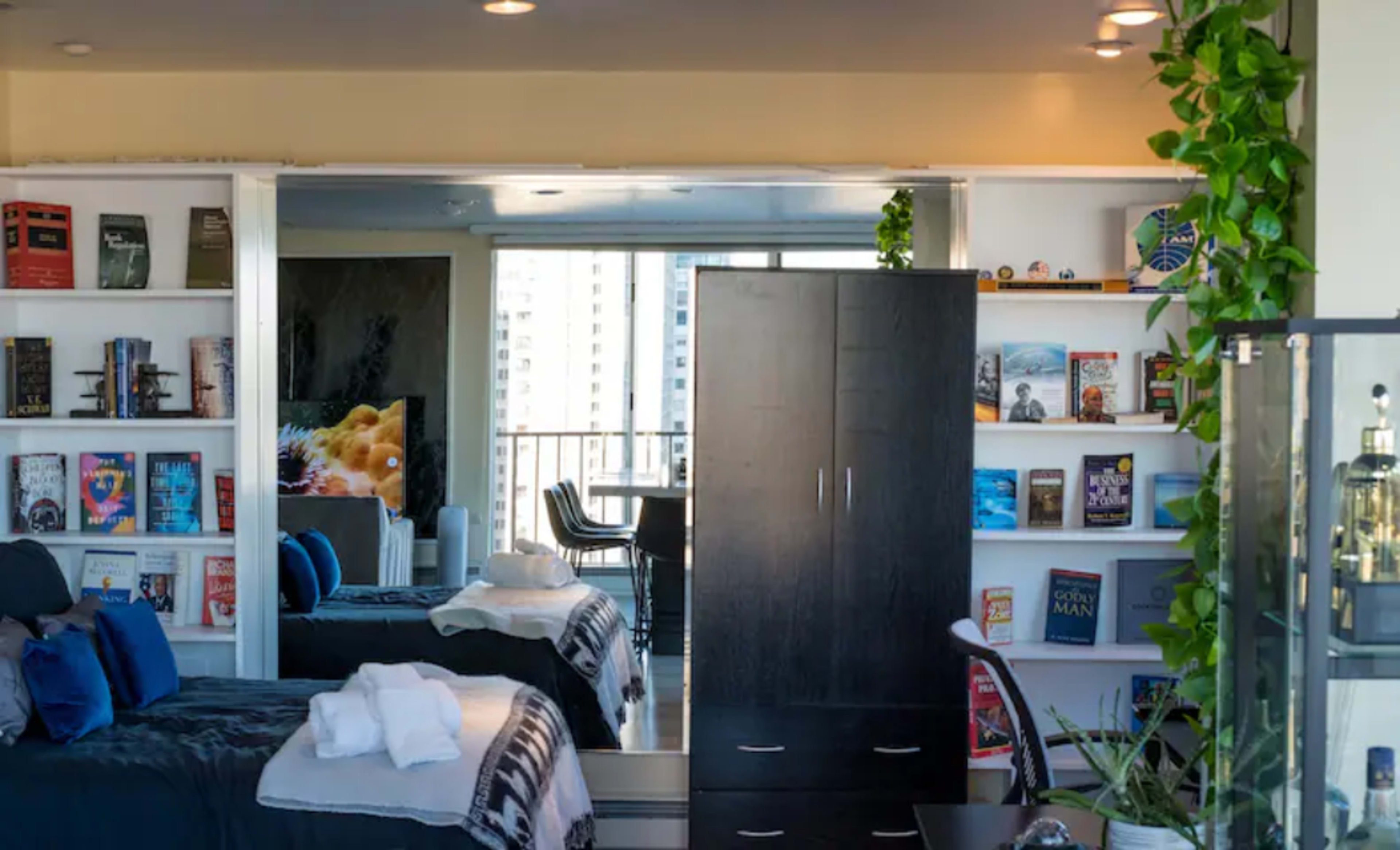 A modern bedroom with a bed, a dark wooden wardrobe, and shelves filled with books, alongside a large window overlooking a cityscape.